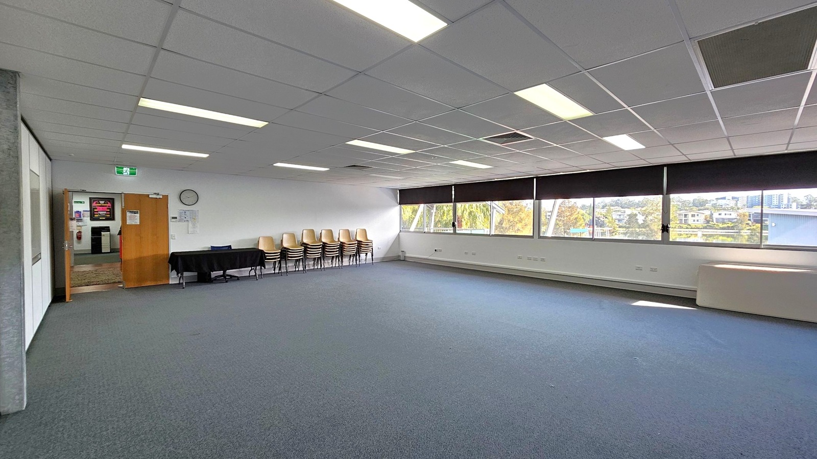 A spacious room with a grey and blue carpeted floor. On the left, there is an open wooden door leading to another area, with a clock and some notices on the wall. A black table with stacked chairs is nearby. The right side features large windows with black blinds, offering a view of trees and buildings outside. The ceiling is fitted with fluorescent lights and ceiling tiles. A spacious room with a grey and blue carpeted floor. On the left, there is an open wooden door leading to another area, with a clock and some notices on the wall. A black table with stacked chairs is nearby. The right side features large windows with black blinds, offering a view of trees and buildings outside. The ceiling is fitted with fluorescent lights and ceiling tiles.
