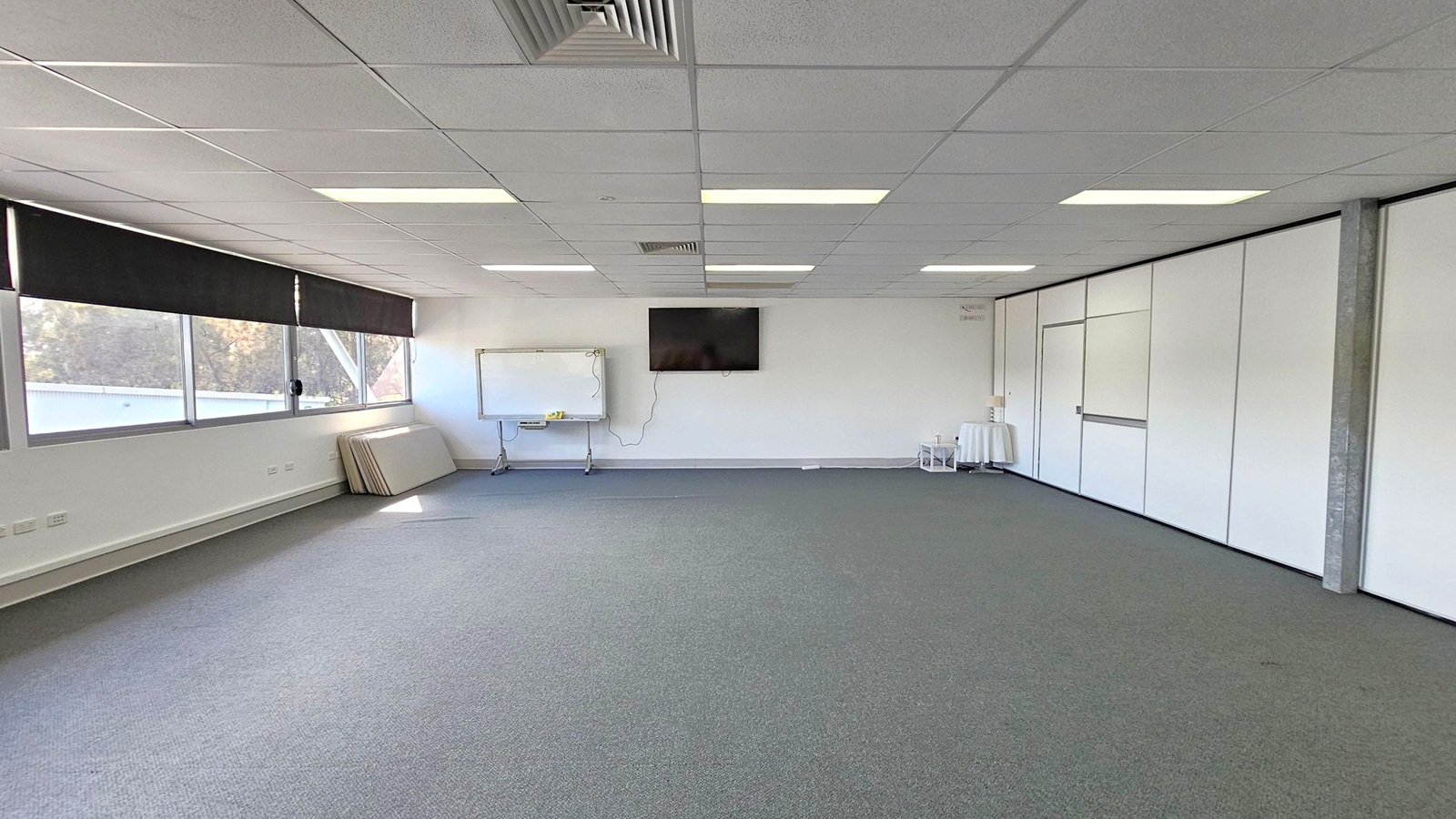 A spacious room with a grey carpeted floor and a white tiled ceiling featuring fluorescent lights. On the left, there are large windows with black blinds, and several folded tables are stacked against the wall. In the centre, a whiteboard and a wall-mounted TV are visible. On the right, there are white partition walls with a door. A small table with a cloth and some items is near the corner. A spacious room with a grey carpeted floor and a white tiled ceiling featuring fluorescent lights. On the left, there are large windows with black blinds, and several folded tables are stacked against the wall. In the centre, a whiteboard and a wall-mounted TV are visible. On the right, there are white partition walls with a door. A small table with a cloth and some items is near the corner.