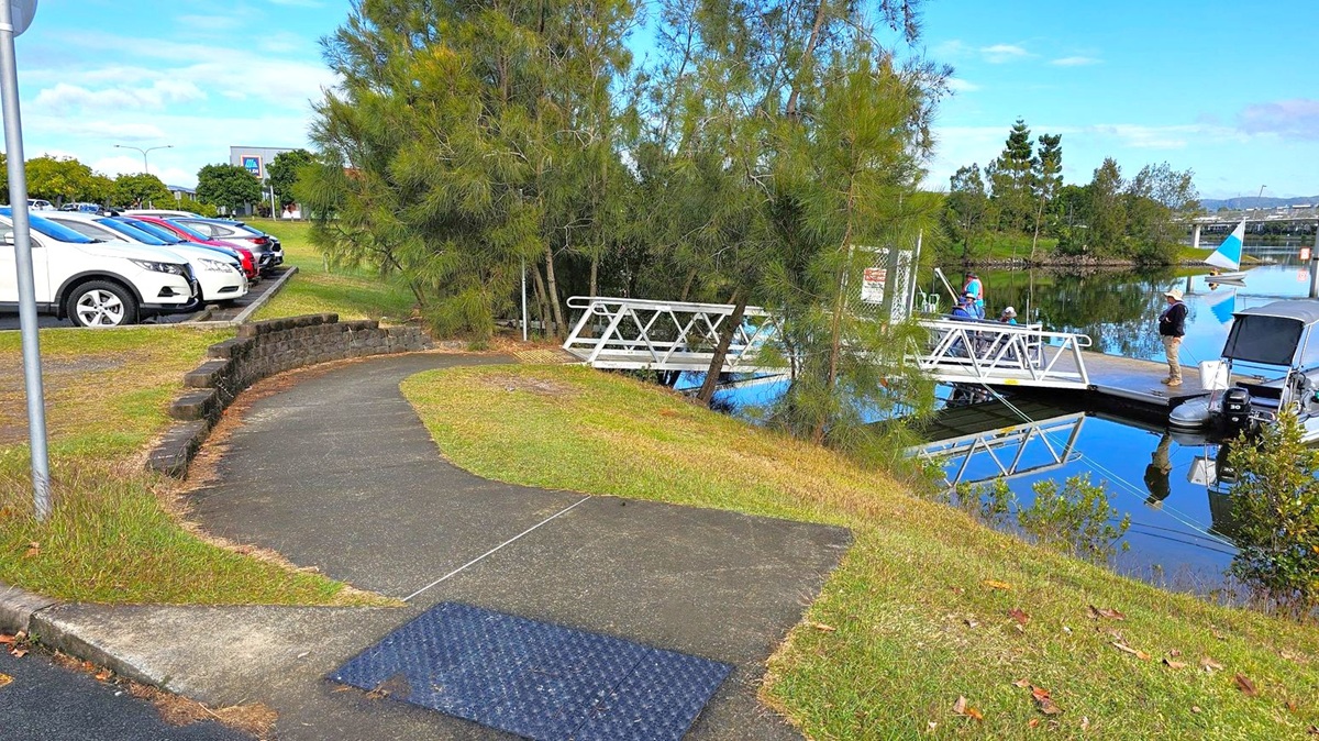 A curved path leads towards a small dock on the right, where a few people are standing near a boat. The dock is surrounded by calm water reflecting the scene. On the left, a row of parked cars is visible, with a grassy area and a low stone wall separating the path from the parking lot. Trees are scattered throughout the area, providing some shade. In the background, a small sailboat is on the water, and a bridge is visible in the distance. A curved path leads towards a small dock on the right, where a few people are standing near a boat. The dock is surrounded by calm water reflecting the scene. On the left, a row of parked cars is visible, with a grassy area and a low stone wall separating the path from the parking lot. Trees are scattered throughout the area, providing some shade. In the background, a small sailboat is on the water, and a bridge is visible in the distance.