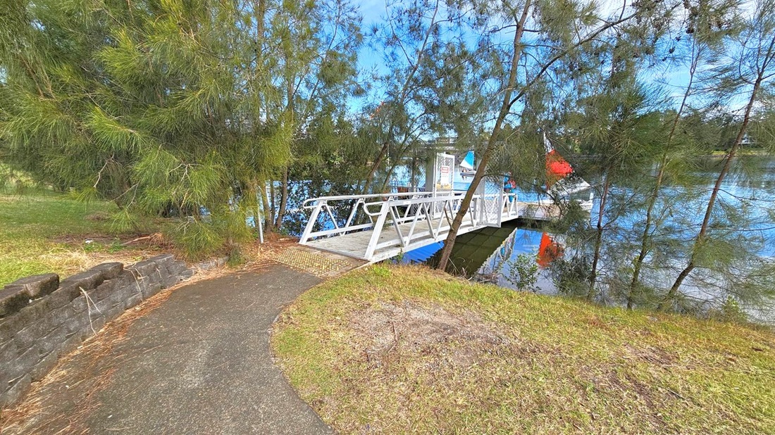 A narrow path leads to a white metal bridge over a body of water. On the left, dense green trees and a low stone wall line the path. The bridge extends to a small dock where a person stands near a sailboat with a red and black sail. The water reflects the trees and the boat. The ground is grassy with patches of dirt. A narrow path leads to a white metal bridge over a body of water. On the left, dense green trees and a low stone wall line the path. The bridge extends to a small dock where a person stands near a sailboat with a red and black sail. The water reflects the trees and the boat. The ground is grassy with patches of dirt.