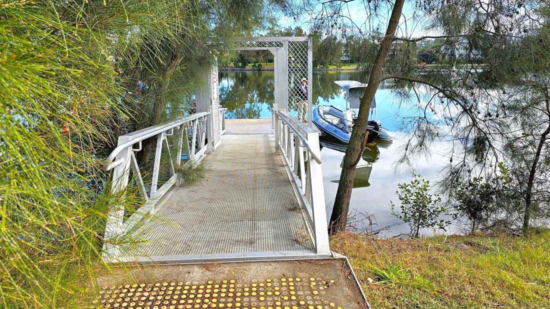 A metal walkway with railings extends towards a small dock over a calm body of water. On the left, dense green foliage partially obscures the view. On the right, a tree with thin branches leans over the water, where a small boat is moored. The background features a reflective water surface with trees lining the opposite bank. The ground near the walkway is grassy with a tactile paving surface at the entrance. A metal walkway with railings extends towards a small dock over a calm body of water. On the left, dense green foliage partially obscures the view. On the right, a tree with thin branches leans over the water, where a small boat is moored. The background features a reflective water surface with trees lining the opposite bank. The ground near the walkway is grassy with a tactile paving surface at the entrance.