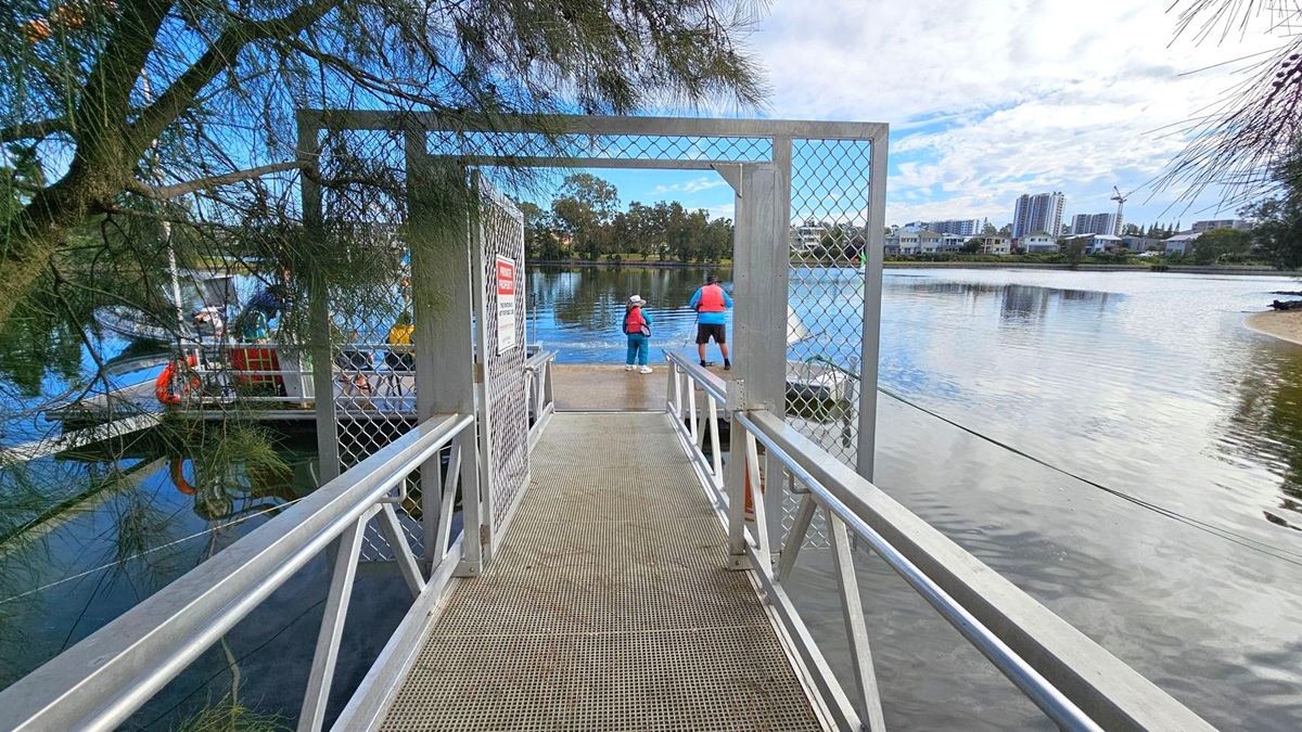 A metal walkway with a mesh gate leads to a dock over a body of water. Two people in life jackets stand at the end of the dock, facing the water. On the left, a tree with dense branches partially obscures the view. In the background, across the water, there are buildings and a crane, indicating an urban area. The water reflects the surroundings, and the sky is partly cloudy. A metal walkway with a mesh gate leads to a dock over a body of water. Two people in life jackets stand at the end of the dock, facing the water. On the left, a tree with dense branches partially obscures the view. In the background, across the water, there are buildings and a crane, indicating an urban area. The water reflects the surroundings, and the sky is partly cloudy.