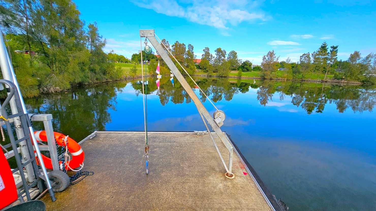 A calm lake with clear reflections of trees and a grassy bank in the background. In the foreground, a concrete platform extends over the water, featuring a metal crane with a pulley system and a hook. To the left, a red and white lifebuoy is attached to a metal structure with wheels. The sky is blue with scattered clouds, and the overall scene is tranquil and still. A calm lake with clear reflections of trees and a grassy bank in the background. In the foreground, a concrete platform extends over the water, featuring a metal crane with a pulley system and a hook. To the left, a red and white lifebuoy is attached to a metal structure with wheels. The sky is blue with scattered clouds, and the overall scene is tranquil and still.