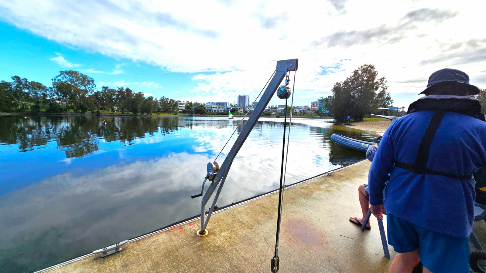 A calm body of water reflects the sky and surrounding trees. On the left, a metal crane with a pulley system is mounted on a concrete surface. In the background, modern buildings are visible across the water. On the right, a person wearing a blue outfit and hat stands near the edge, with another person seated nearby. A small inflatable boat is partially visible near the shore. Trees line the far bank, and the sky is partly cloudy. A calm body of water reflects the sky and surrounding trees. On the left, a metal crane with a pulley system is mounted on a concrete surface. In the background, modern buildings are visible across the water. On the right, a person wearing a blue outfit and hat stands near the edge, with another person seated nearby. A small inflatable boat is partially visible near the shore. Trees line the far bank, and the sky is partly cloudy.