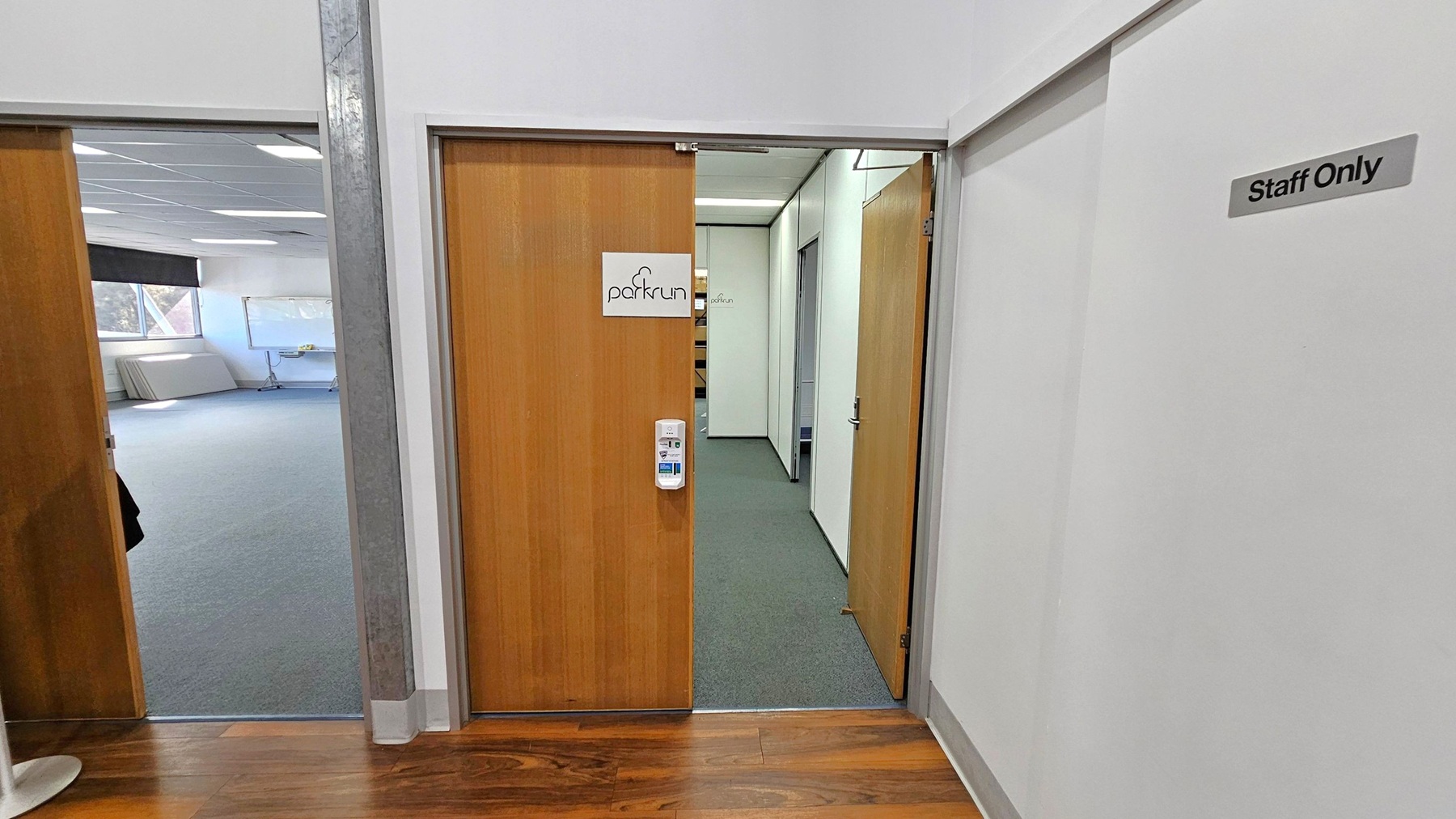 A hallway with two wooden doors. The door on the right has a sign reading "parkrun" and a hand sanitiser dispenser mounted on it. The wall next to it has a "Staff Only" sign. The door on the left opens into a room with grey carpet, a large window, and a whiteboard. The floor in the hallway is wooden, and the ceiling has recessed lighting. A hallway with two wooden doors. The door on the right has a sign reading "parkrun" and a hand sanitiser dispenser mounted on it. The wall next to it has a "Staff Only" sign. The door on the left opens into a room with grey carpet, a large window, and a whiteboard. The floor in the hallway is wooden, and the ceiling has recessed lighting.