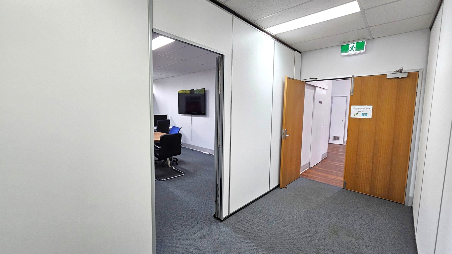 A corridor with grey carpeted flooring and white walls. On the right, a wooden door is partially open, revealing a room with wooden flooring. Above the door, an emergency exit sign is visible. On the left, an open doorway leads to a meeting room with a table, chairs, and a wall-mounted screen. The ceiling has fluorescent lighting. A corridor with grey carpeted flooring and white walls. On the right, a wooden door is partially open, revealing a room with wooden flooring. Above the door, an emergency exit sign is visible. On the left, an open doorway leads to a meeting room with a table, chairs, and a wall-mounted screen. The ceiling has fluorescent lighting.