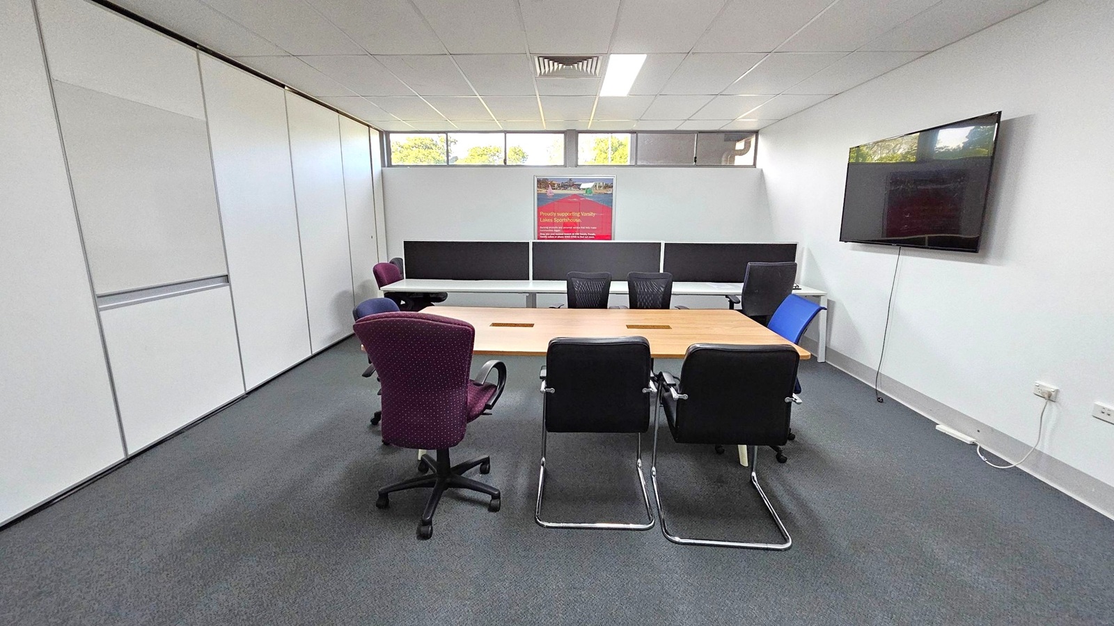 A conference room with a rectangular wooden table in the centre, surrounded by various chairs, including black, blue, and maroon ones. On the right wall, a large flat-screen TV is mounted. The back wall features a poster and windows above, allowing natural light. The left wall is lined with white panels. The floor is carpeted in grey, and the ceiling is fitted with white tiles and fluorescent lights. A conference room with a rectangular wooden table in the centre, surrounded by various chairs, including black, blue, and maroon ones. On the right wall, a large flat-screen TV is mounted. The back wall features a poster and windows above, allowing natural light. The left wall is lined with white panels. The floor is carpeted in grey, and the ceiling is fitted with white tiles and fluorescent lights.