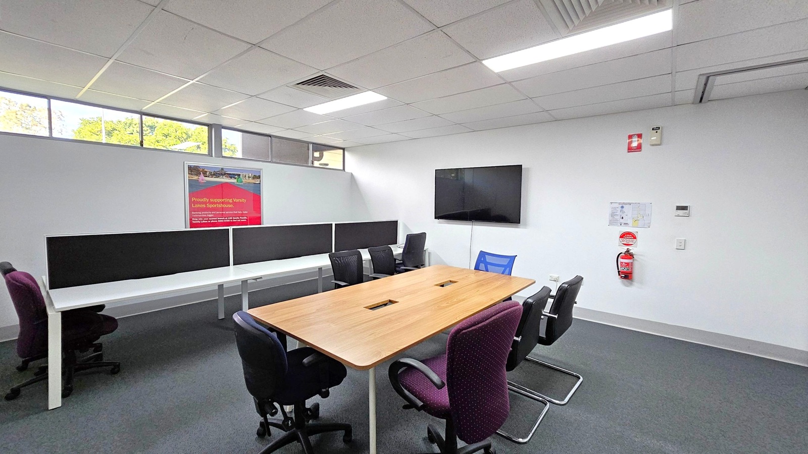 A modern office space with a rectangular wooden table in the centre, surrounded by various office chairs. On the left, there are white desks with black dividers and additional chairs. A red poster is mounted on the wall above the desks. On the right, a flat-screen TV is mounted on the wall, with a fire extinguisher and a small map below it. The ceiling is fitted with fluorescent lights and air vents, and the floor is carpeted in grey. High windows allow natural light in. A modern office space with a rectangular wooden table in the centre, surrounded by various office chairs. On the left, there are white desks with black dividers and additional chairs. A red poster is mounted on the wall above the desks. On the right, a flat-screen TV is mounted on the wall, with a fire extinguisher and a small map below it. The ceiling is fitted with fluorescent lights and air vents, and the floor is carpeted in grey. High windows allow natural light in.