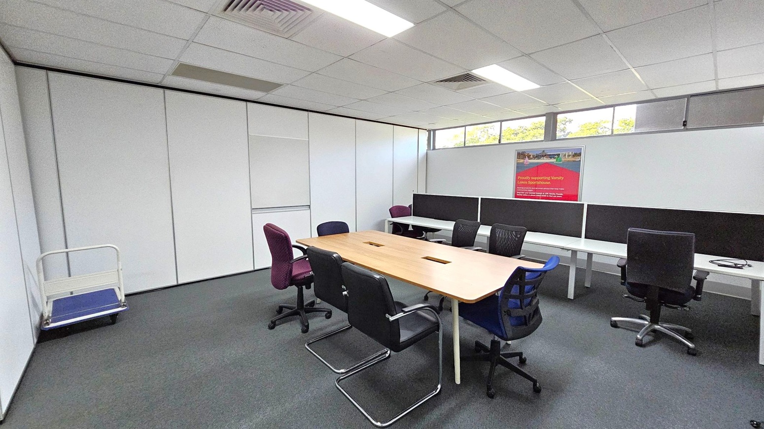 A conference room with a long wooden table in the centre surrounded by various office chairs, including black, purple, and blue ones. On the right, there are white desks with black dividers and additional chairs. A poster is mounted on the wall above the desks. The left side has a small trolley against a white partition wall. The ceiling is fitted with fluorescent lights and air vents, and the floor is carpeted in grey. High windows near the ceiling let in natural light. A conference room with a long wooden table in the centre surrounded by various office chairs, including black, purple, and blue ones. On the right, there are white desks with black dividers and additional chairs. A poster is mounted on the wall above the desks. The left side has a small trolley against a white partition wall. The ceiling is fitted with fluorescent lights and air vents, and the floor is carpeted in grey. High windows near the ceiling let in natural light.