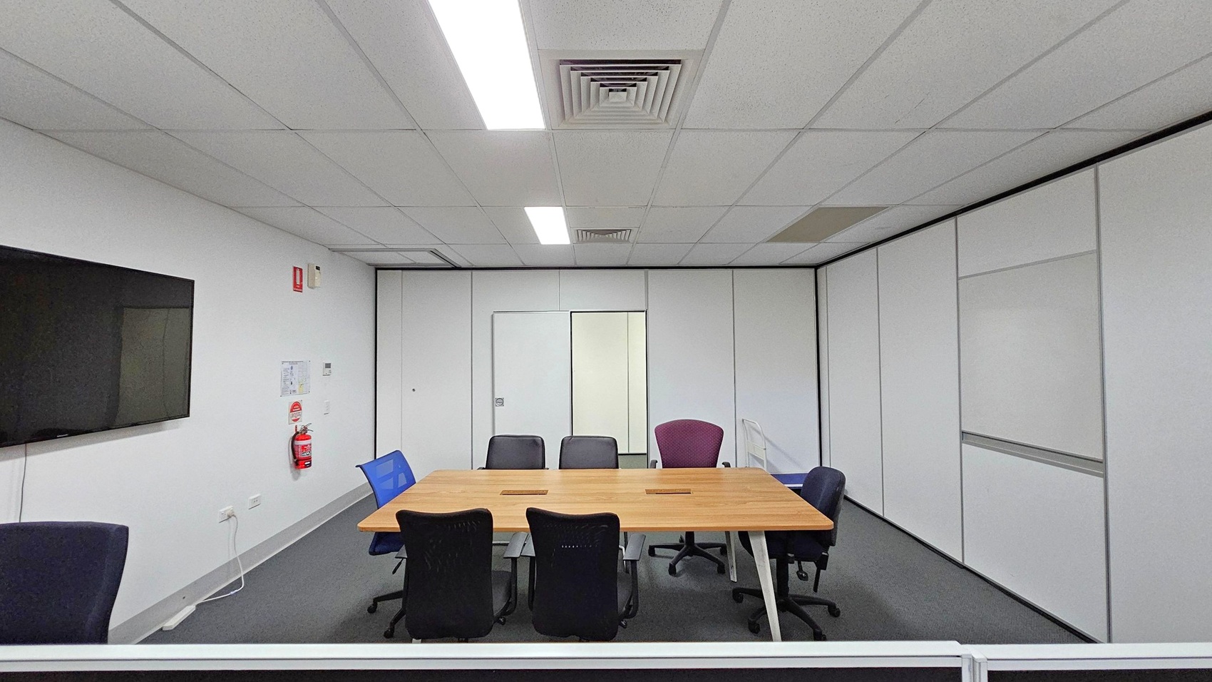 A conference room with a rectangular wooden table in the centre, surrounded by various office chairs. On the left wall, a large flat-screen TV is mounted, with a fire extinguisher and some notices below it. The walls are white with a door in the centre. The ceiling has fluorescent lights and air vents, and the floor is carpeted in grey. A conference room with a rectangular wooden table in the centre, surrounded by various office chairs. On the left wall, a large flat-screen TV is mounted, with a fire extinguisher and some notices below it. The walls are white with a door in the centre. The ceiling has fluorescent lights and air vents, and the floor is carpeted in grey.