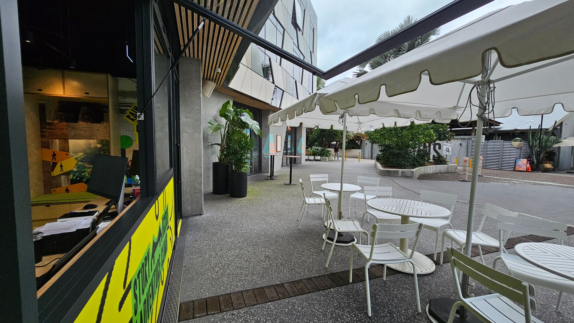 On the left, there's a window with a yellow and green sign, and inside, a desk with a computer monitor and papers. The ceiling has wooden slats. In the middle, there are white tables and chairs under a white canopy. On the right, a pathway leads to a building with a modern facade. Potted plants are near the entrance, and a few people are walking in the distance. The ground is paved with a mix of concrete and brick.