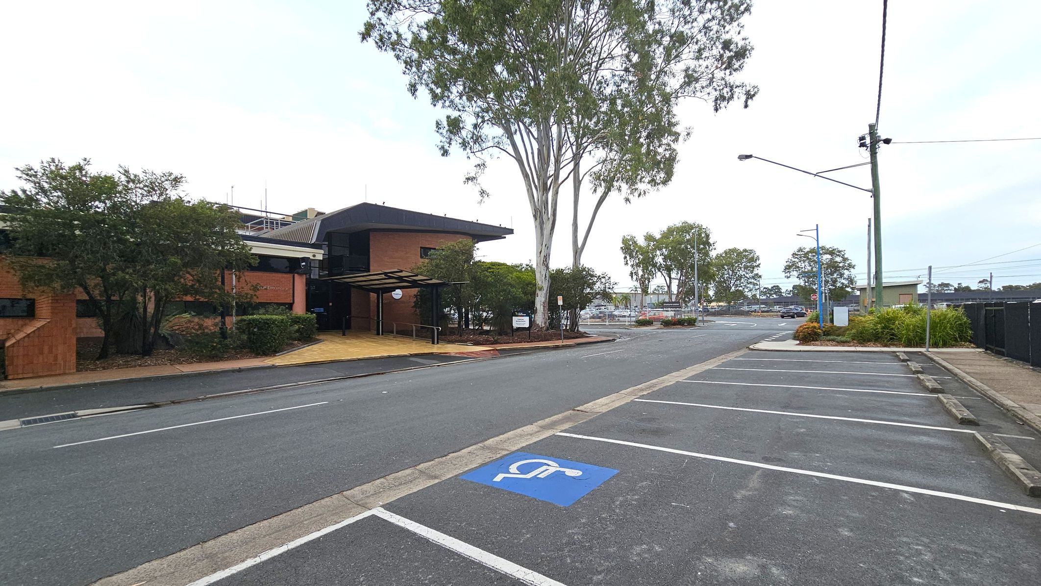 A street scene with a brick building on the left, featuring a covered entrance and surrounded by trees and shrubs. A large tree stands near the building. On the right, there is a parking area with marked spaces, including a designated disabled parking spot. The background shows more trees, a fence, and a few buildings. A streetlight and utility pole are visible near the parking area. The road is empty, and the pavement is bordered by a narrow footpath. A street scene with a brick building on the left, featuring a covered entrance and surrounded by trees and shrubs. A large tree stands near the building. On the right, there is a parking area with marked spaces, including a designated disabled parking spot. The background shows more trees, a fence, and a few buildings. A streetlight and utility pole are visible near the parking area. The road is empty, and the pavement is bordered by a narrow footpath.