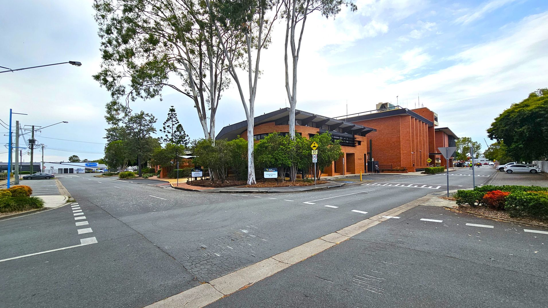 A street intersection with a brick building on the right, featuring a modern design and surrounded by trees. The road has marked pedestrian crossings and a give-way sign. On the left, there are more trees and a few parked cars. The background shows additional buildings and a clear sky. The foreground has a paved road with visible lane markings. A street intersection with a brick building on the right, featuring a modern design and surrounded by trees. The road has marked pedestrian crossings and a give-way sign. On the left, there are more trees and a few parked cars. The background shows additional buildings and a clear sky. The foreground has a paved road with visible lane markings.