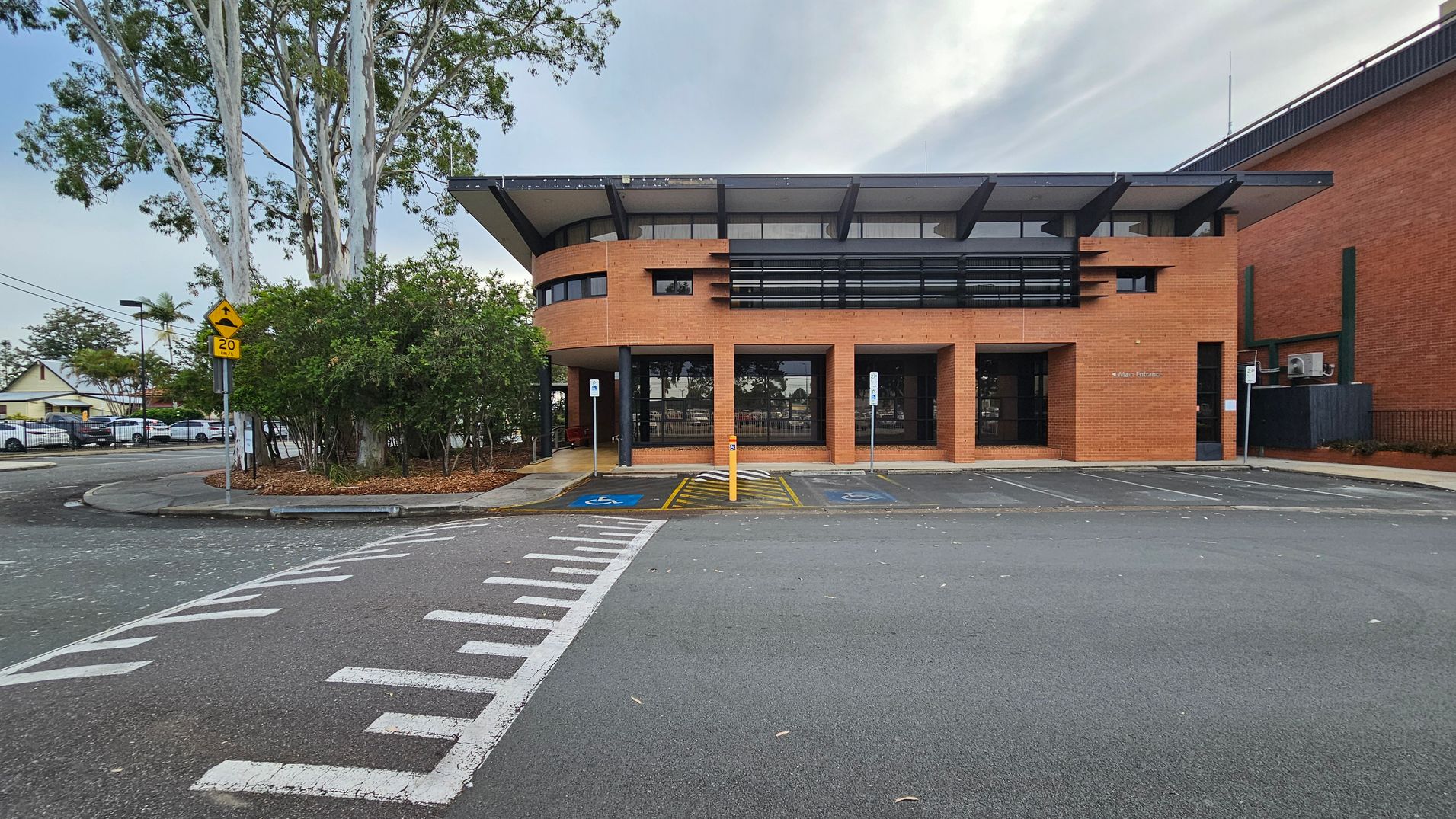 A brick building with a modern design features large windows and a flat roof with overhangs. In front, there are two marked disabled parking spaces and a pedestrian crossing. To the left, a cluster of trees and shrubs is visible, along with a yellow road sign indicating a speed limit of 20. The road curves around the building, and a few parked cars are seen in the background. The pavement is grey with white markings. A brick building with a modern design features large windows and a flat roof with overhangs. In front, there are two marked disabled parking spaces and a pedestrian crossing. To the left, a cluster of trees and shrubs is visible, along with a yellow road sign indicating a speed limit of 20. The road curves around the building, and a few parked cars are seen in the background. The pavement is grey with white markings.