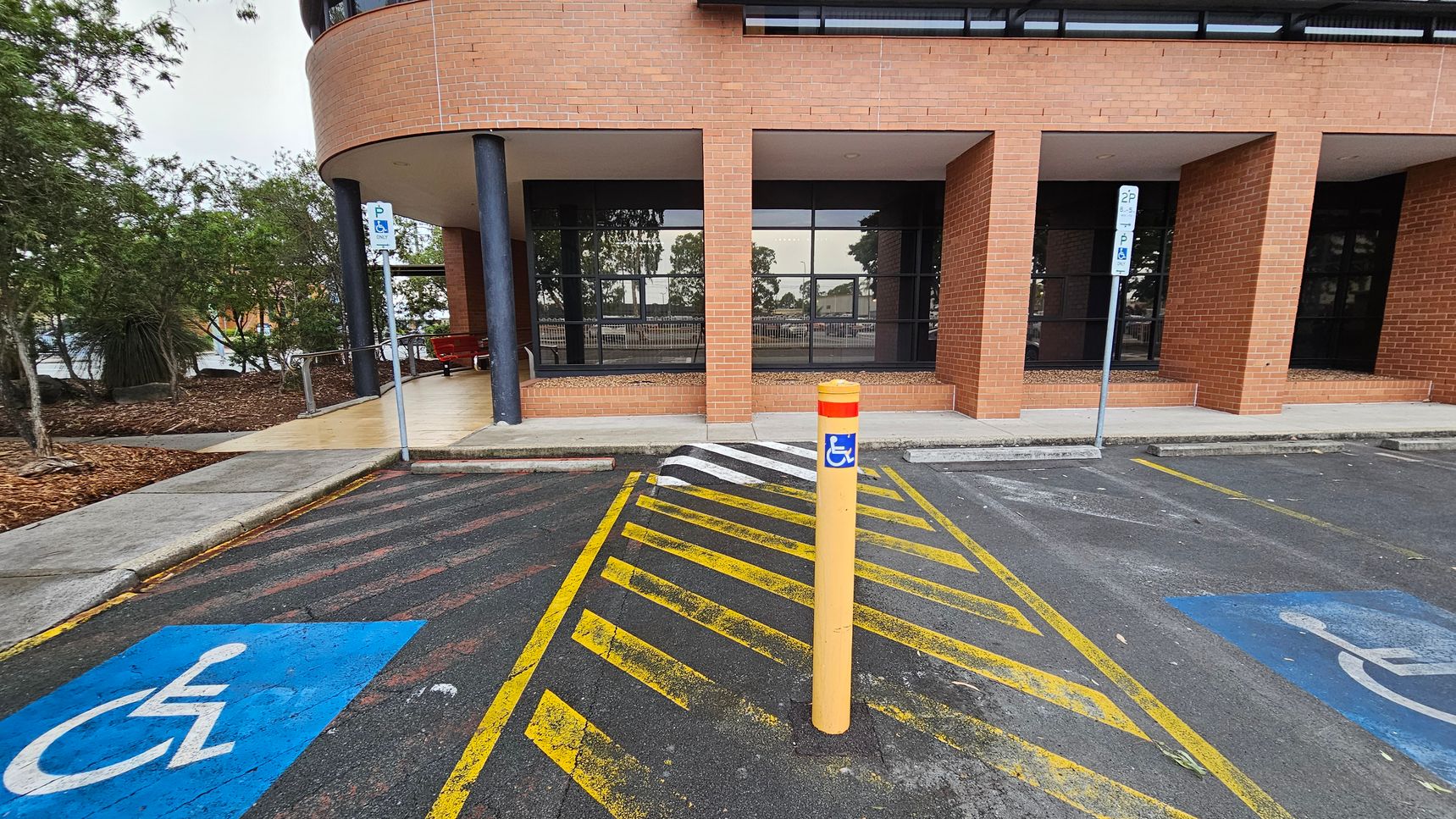 A parking area with two marked disabled parking spaces on either side, featuring blue wheelchair symbols. In the centre, a yellow bollard with a blue disabled sign stands on a yellow-striped zone. The background shows a brick building with large windows and a covered walkway supported by black columns. To the left, there are trees and a landscaped area with mulch. The ground is asphalt, and the ceiling of the walkway is light-coloured. A parking area with two marked disabled parking spaces on either side, featuring blue wheelchair symbols. In the centre, a yellow bollard with a blue disabled sign stands on a yellow-striped zone. The background shows a brick building with large windows and a covered walkway supported by black columns. To the left, there are trees and a landscaped area with mulch. The ground is asphalt, and the ceiling of the walkway is light-coloured.