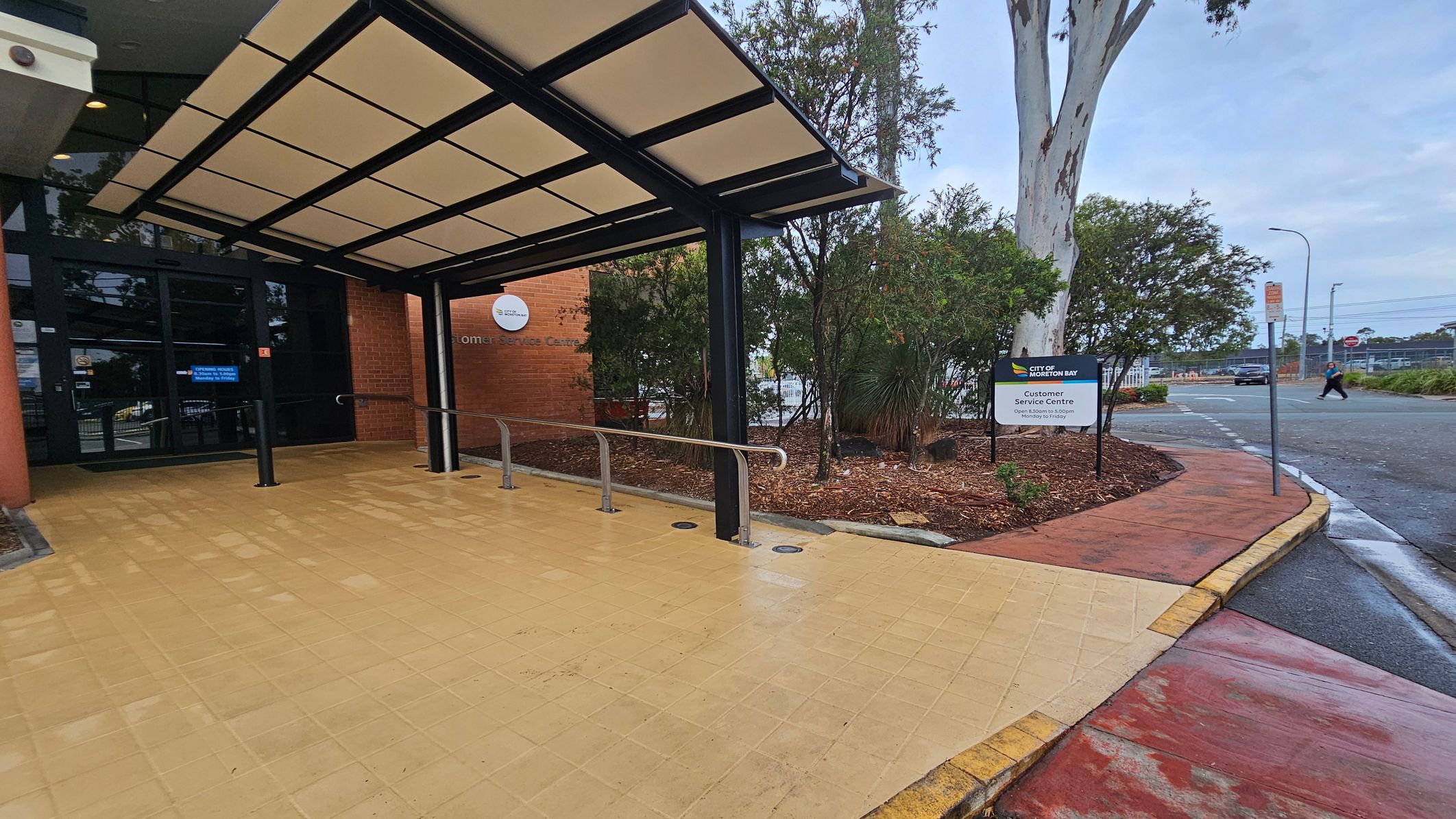 Entrance to a building with a covered walkway on the left, featuring a glass door and a sign indicating a customer service centre. The floor is tiled in a light yellow colour. On the right, there is a landscaped area with bushes and a tree, alongside a sign for the City of Kwinana. A pavement leads to a road where a person is crossing. The ceiling of the walkway is structured with a black frame and light panels. Entrance to a building with a covered walkway on the left, featuring a glass door and a sign indicating a customer service centre. The floor is tiled in a light yellow colour. On the right, there is a landscaped area with bushes and a tree, alongside a sign for the City of Kwinana. A pavement leads to a road where a person is crossing. The ceiling of the walkway is structured with a black frame and light panels.