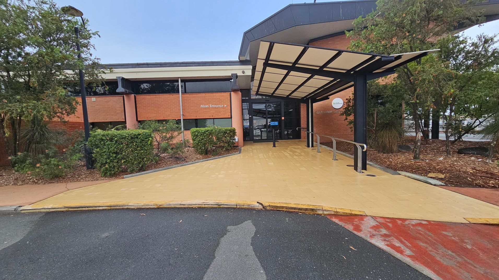 A building entrance with a covered walkway leading to glass doors. The structure has a brick facade with a sign indicating the main entrance. On the left, there are bushes and a lamp post, while on the right, more trees and shrubs are visible. The ground is paved with a yellowish surface leading from the road, which is partially visible in the foreground. The roof of the entrance is supported by black metal beams. A building entrance with a covered walkway leading to glass doors. The structure has a brick facade with a sign indicating the main entrance. On the left, there are bushes and a lamp post, while on the right, more trees and shrubs are visible. The ground is paved with a yellowish surface leading from the road, which is partially visible in the foreground. The roof of the entrance is supported by black metal beams.