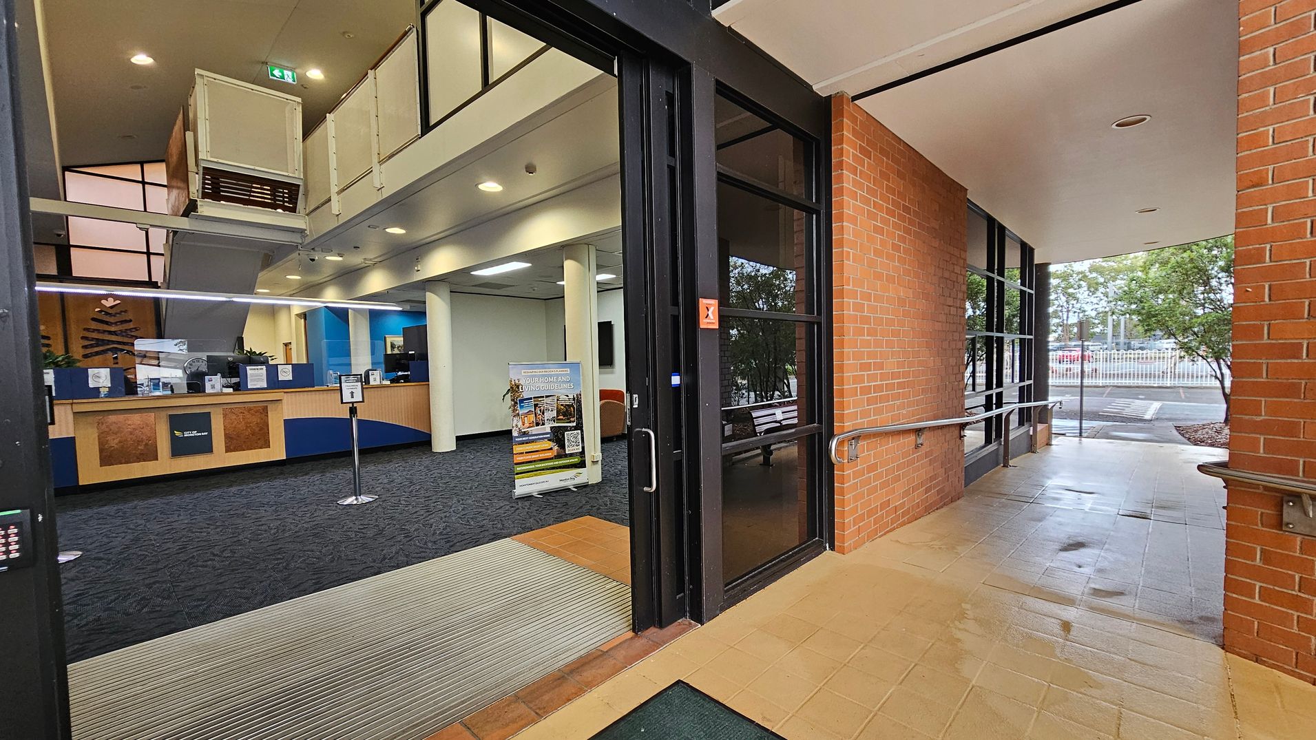 Entrance to a building with a glass door on the right, leading to a tiled corridor with brick walls and metal handrails. Inside, a reception area with a wooden counter and blue accents is visible. A sign with information stands on the carpeted floor. The ceiling has recessed lighting, and a staircase is visible in the background. Outside, trees and a parking area can be seen through the glass. Entrance to a building with a glass door on the right, leading to a tiled corridor with brick walls and metal handrails. Inside, a reception area with a wooden counter and blue accents is visible. A sign with information stands on the carpeted floor. The ceiling has recessed lighting, and a staircase is visible in the background. Outside, trees and a parking area can be seen through the glass.