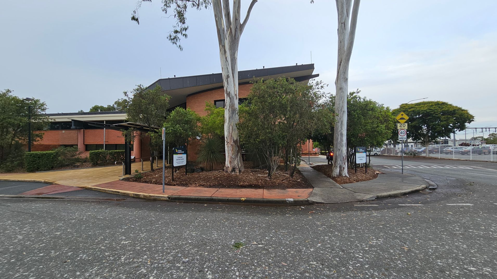A brick building with a sloped roof is in the centre, surrounded by trees and shrubs. A large tree stands prominently in front. On the right, a road with speed limit signs curves around the building. A footpath runs alongside the road, bordered by landscaped garden beds. In the background, a fence and parked cars are visible. The ground is paved with a mix of asphalt and concrete. A brick building with a sloped roof is in the centre, surrounded by trees and shrubs. A large tree stands prominently in front. On the right, a road with speed limit signs curves around the building. A footpath runs alongside the road, bordered by landscaped garden beds. In the background, a fence and parked cars are visible. The ground is paved with a mix of asphalt and concrete.