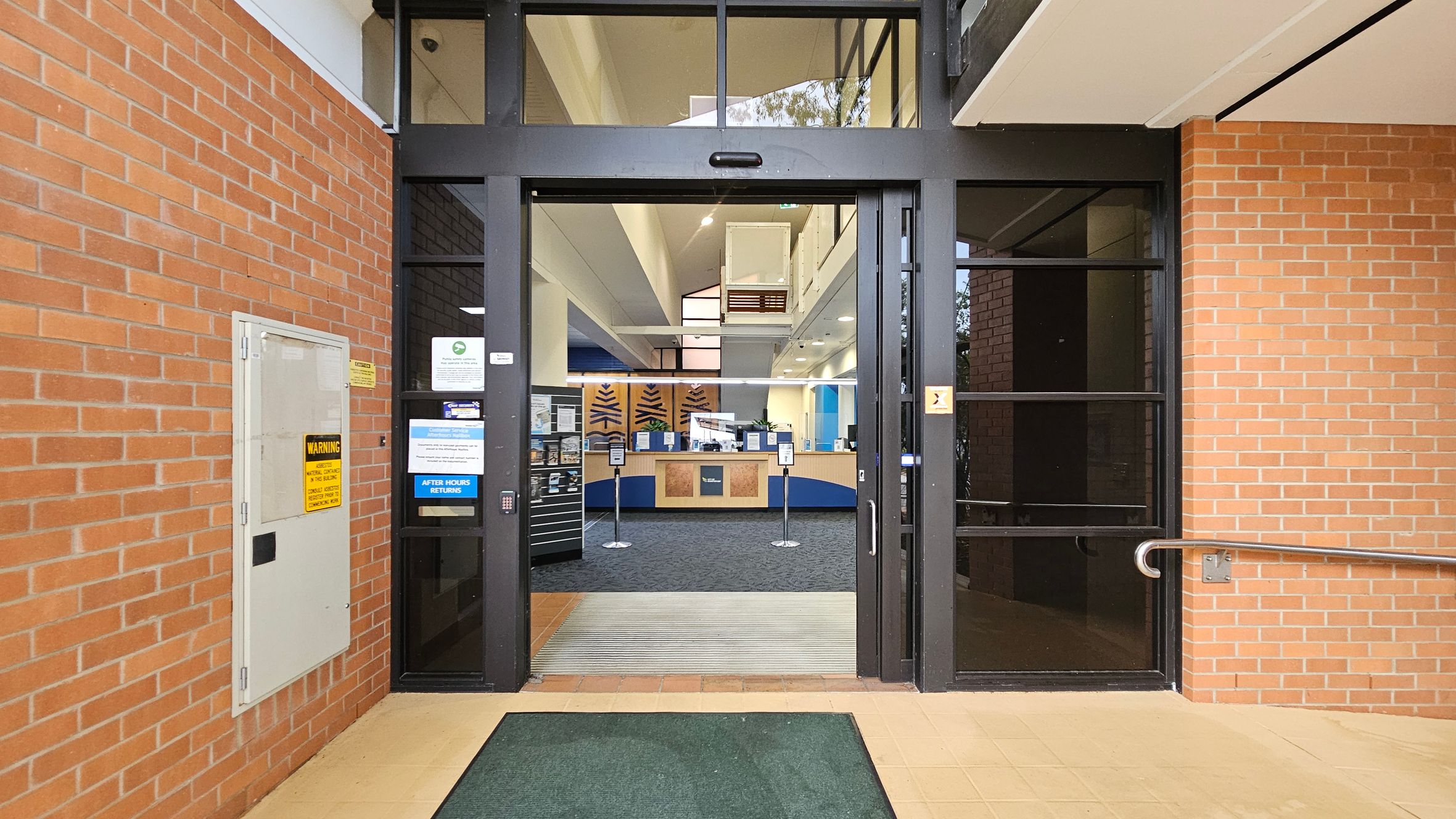 Entrance to a building with a glass door framed in black, flanked by red brick walls. On the left, a white panel with warning signs is mounted on the brick wall. Inside, a reception area with a counter and signage is visible. The floor outside is tiled with a green mat, and inside, the floor is carpeted. The ceiling is high with recessed lighting. A metal handrail is attached to the right wall. Entrance to a building with a glass door framed in black, flanked by red brick walls. On the left, a white panel with warning signs is mounted on the brick wall. Inside, a reception area with a counter and signage is visible. The floor outside is tiled with a green mat, and inside, the floor is carpeted. The ceiling is high with recessed lighting. A metal handrail is attached to the right wall.
