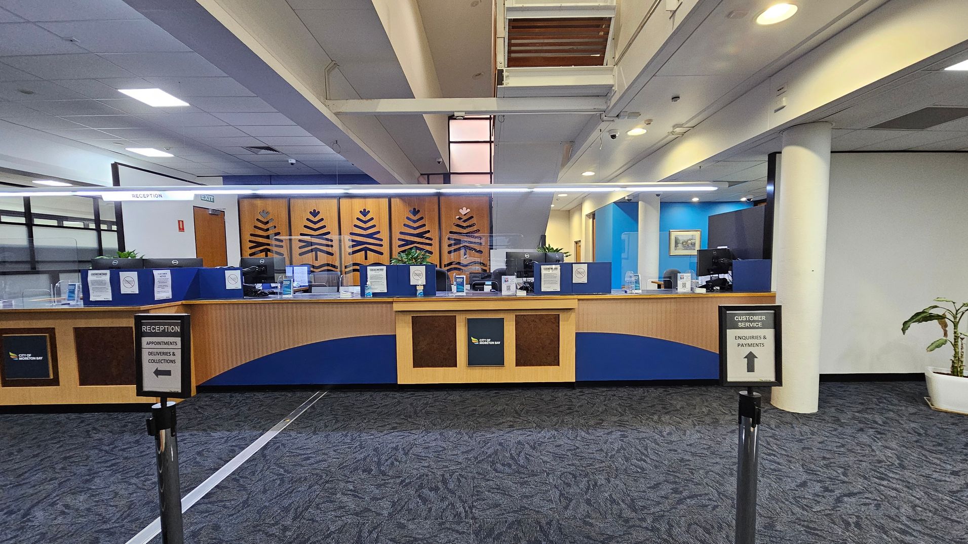Reception area with a long wooden counter featuring blue accents and decorative panels. Behind the counter are computer monitors and signs. Two signs in the foreground direct visitors to reception and customer service. The floor is carpeted in a dark pattern, and the ceiling has recessed lighting. A potted plant is visible on the right, and the walls are painted in blue and white tones. An exit sign is above the counter. Reception area with a long wooden counter featuring blue accents and decorative panels. Behind the counter are computer monitors and signs. Two signs in the foreground direct visitors to reception and customer service. The floor is carpeted in a dark pattern, and the ceiling has recessed lighting. A potted plant is visible on the right, and the walls are painted in blue and white tones. An exit sign is above the counter.