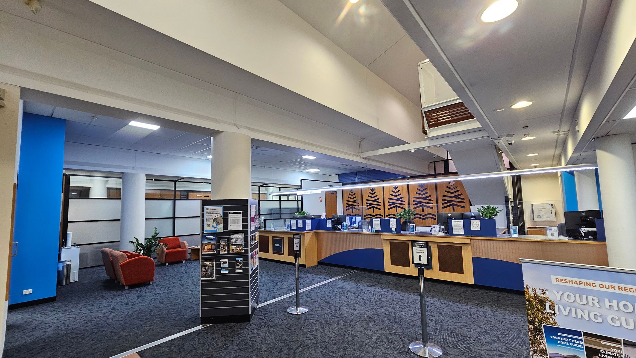 A spacious interior with a reception area on the right, featuring a wooden counter with blue accents and decorative panels. Potted plants are placed on the counter. In the foreground, a sign and a brochure stand are visible. On the left, there are red armchairs and a small table near a blue wall. The floor is carpeted in a dark pattern, and the ceiling has recessed lighting. Large windows with frosted glass panels are in the background. A spacious interior with a reception area on the right, featuring a wooden counter with blue accents and decorative panels. Potted plants are placed on the counter. In the foreground, a sign and a brochure stand are visible. On the left, there are red armchairs and a small table near a blue wall. The floor is carpeted in a dark pattern, and the ceiling has recessed lighting. Large windows with frosted glass panels are in the background.