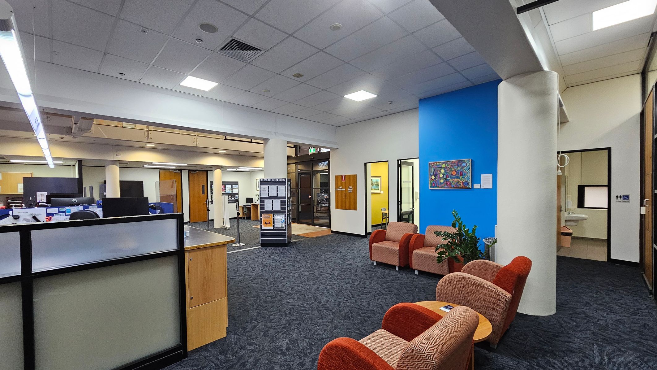 A spacious office area with a blue accent wall on the right, featuring a colourful artwork. In front of the wall are several cushioned chairs and a small table. A plant is placed nearby. On the left, there are workstations with computers and a partitioned desk. The floor is carpeted in a dark pattern, and the ceiling has recessed lighting. A public notice board stands near the centre, and there are doors leading to other rooms. A spacious office area with a blue accent wall on the right, featuring a colourful artwork. In front of the wall are several cushioned chairs and a small table. A plant is placed nearby. On the left, there are workstations with computers and a partitioned desk. The floor is carpeted in a dark pattern, and the ceiling has recessed lighting. A public notice board stands near the centre, and there are doors leading to other rooms.