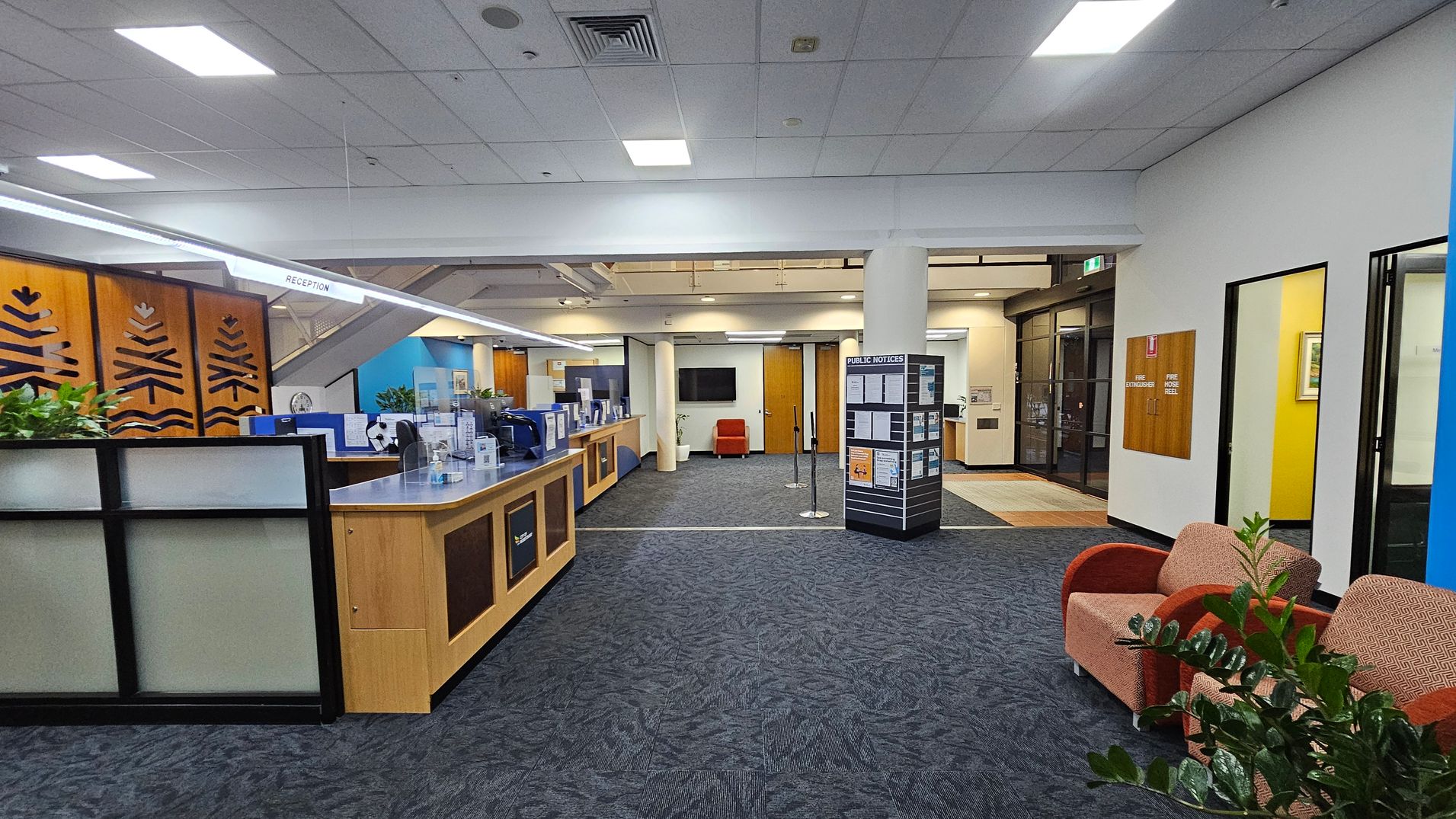 Reception area with a wooden counter on the left, featuring decorative panels and potted plants. A sign above reads "RECEPTION." In the centre, a display board with public notices stands on a carpeted floor. To the right, two upholstered chairs are placed near a plant. The background includes a wall-mounted TV and wooden doors. The ceiling has recessed lighting, and the floor is covered with patterned carpet tiles. Reception area with a wooden counter on the left, featuring decorative panels and potted plants. A sign above reads "RECEPTION." In the centre, a display board with public notices stands on a carpeted floor. To the right, two upholstered chairs are placed near a plant. The background includes a wall-mounted TV and wooden doors. The ceiling has recessed lighting, and the floor is covered with patterned carpet tiles.
