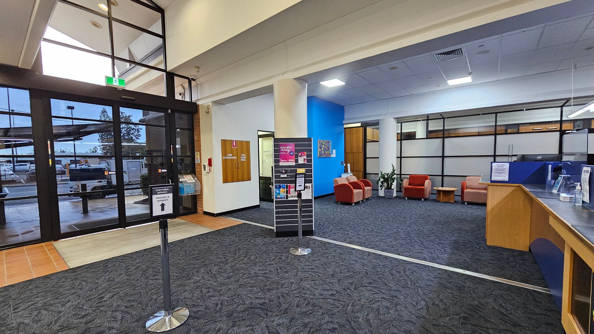 Entrance area of a public building with large glass doors on the left, leading outside. A sign directs to customer service. In the centre, a display stand holds brochures. To the right, a waiting area with red and beige chairs, a small table, and potted plants. The floor is carpeted in dark grey, and the ceiling is fitted with lights and air vents. A wooden counter with a clear screen is on the far right. Entrance area of a public building with large glass doors on the left, leading outside. A sign directs to customer service. In the centre, a display stand holds brochures. To the right, a waiting area with red and beige chairs, a small table, and potted plants. The floor is carpeted in dark grey, and the ceiling is fitted with lights and air vents. A wooden counter with a clear screen is on the far right.