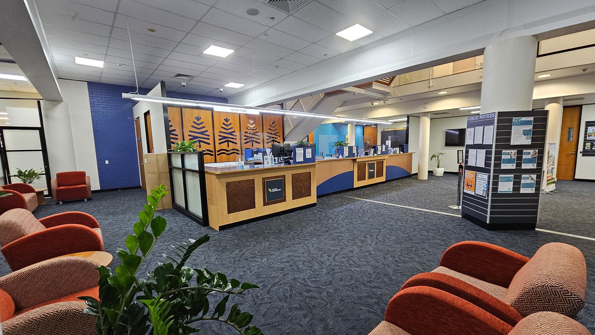 A spacious interior with a reception desk in the centre, featuring wood panelling and decorative designs. To the left, there are several orange upholstered chairs and green plants. On the right, a board displays public notices. The floor is carpeted in a dark blue pattern, and the ceiling is white with recessed lighting. In the background, there are glass doors and a staircase leading to an upper level. A spacious interior with a reception desk in the centre, featuring wood panelling and decorative designs. To the left, there are several orange upholstered chairs and green plants. On the right, a board displays public notices. The floor is carpeted in a dark blue pattern, and the ceiling is white with recessed lighting. In the background, there are glass doors and a staircase leading to an upper level.