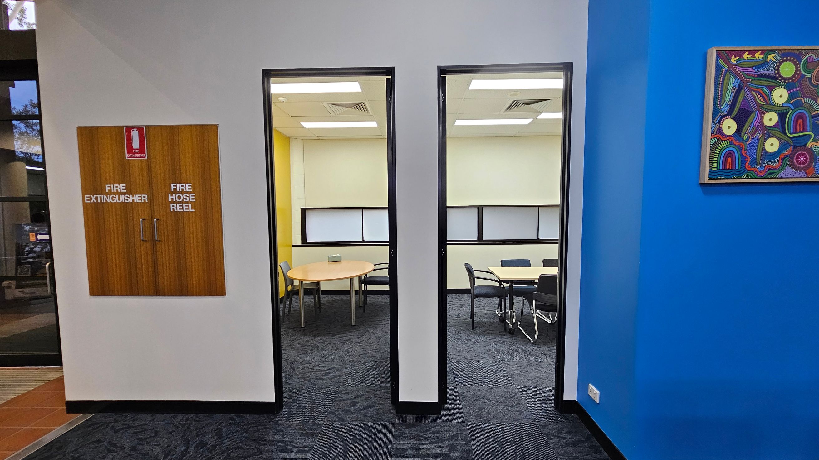 A hallway with a grey wall in the centre featuring a wooden cabinet labelled "Fire Extinguisher" and "Fire Hose Reel". Two doorways lead to rooms with tables and chairs. The left room has a yellow wall, while the right room has a light-coloured wall. On the right, a bright blue wall displays a colourful abstract painting. The floor is carpeted in a dark pattern, and the ceiling has white panels with recessed lighting. A hallway with a grey wall in the centre featuring a wooden cabinet labelled "Fire Extinguisher" and "Fire Hose Reel". Two doorways lead to rooms with tables and chairs. The left room has a yellow wall, while the right room has a light-coloured wall. On the right, a bright blue wall displays a colourful abstract painting. The floor is carpeted in a dark pattern, and the ceiling has white panels with recessed lighting.