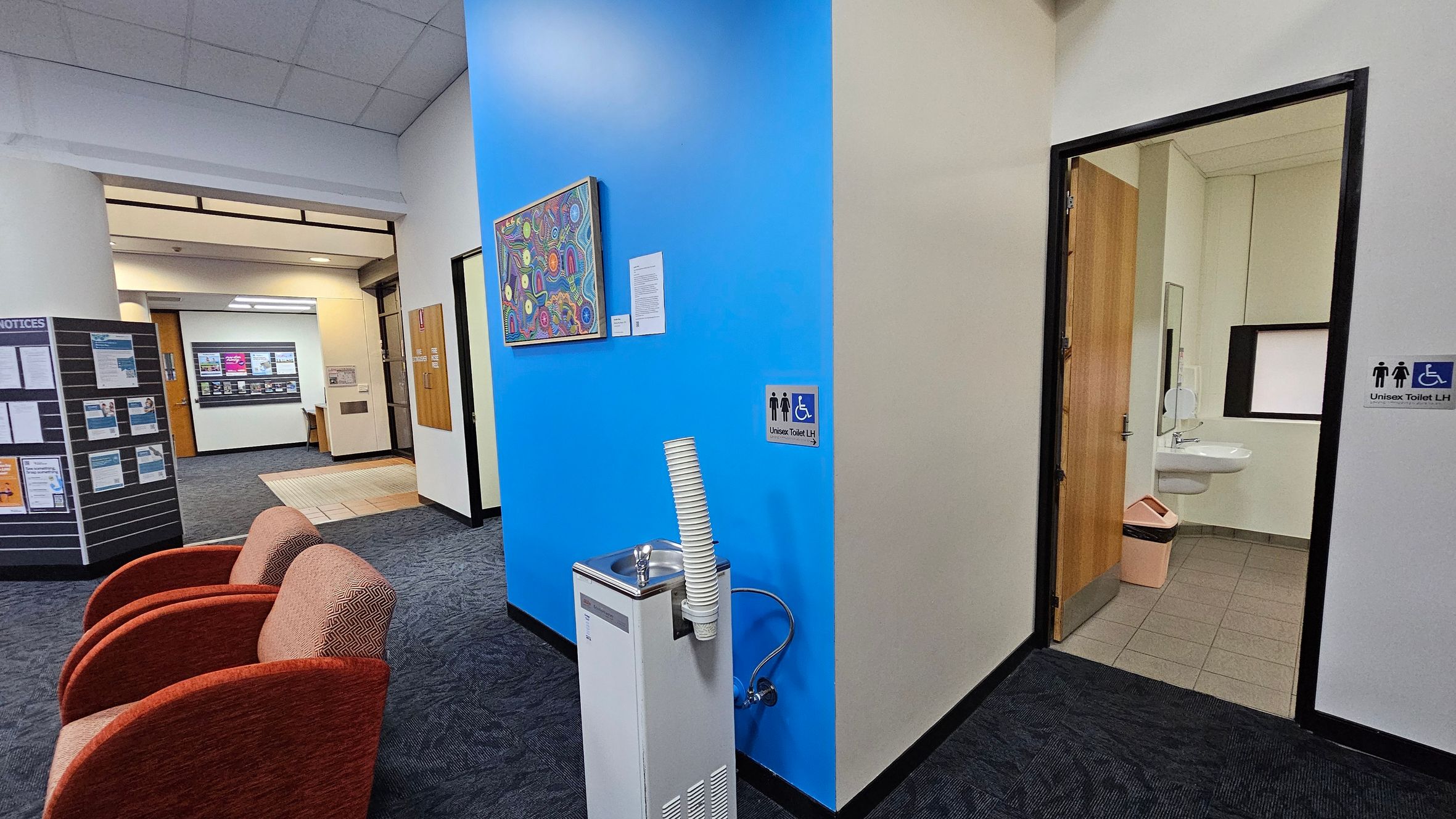 A hallway with a blue accent wall in the centre, featuring a colourful artwork. Below the artwork is a water fountain with stacked cups. To the right, an open door leads to a unisex accessible toilet, visible with a sink and bin. On the left, two red chairs are placed on a dark carpeted floor. Further left, a notice board displays various papers. In the background, a corridor with a light-coloured floor and a wooden door is visible. The ceiling is tiled. A hallway with a blue accent wall in the centre, featuring a colourful artwork. Below the artwork is a water fountain with stacked cups. To the right, an open door leads to a unisex accessible toilet, visible with a sink and bin. On the left, two red chairs are placed on a dark carpeted floor. Further left, a notice board displays various papers. In the background, a corridor with a light-coloured floor and a wooden door is visible. The ceiling is tiled.