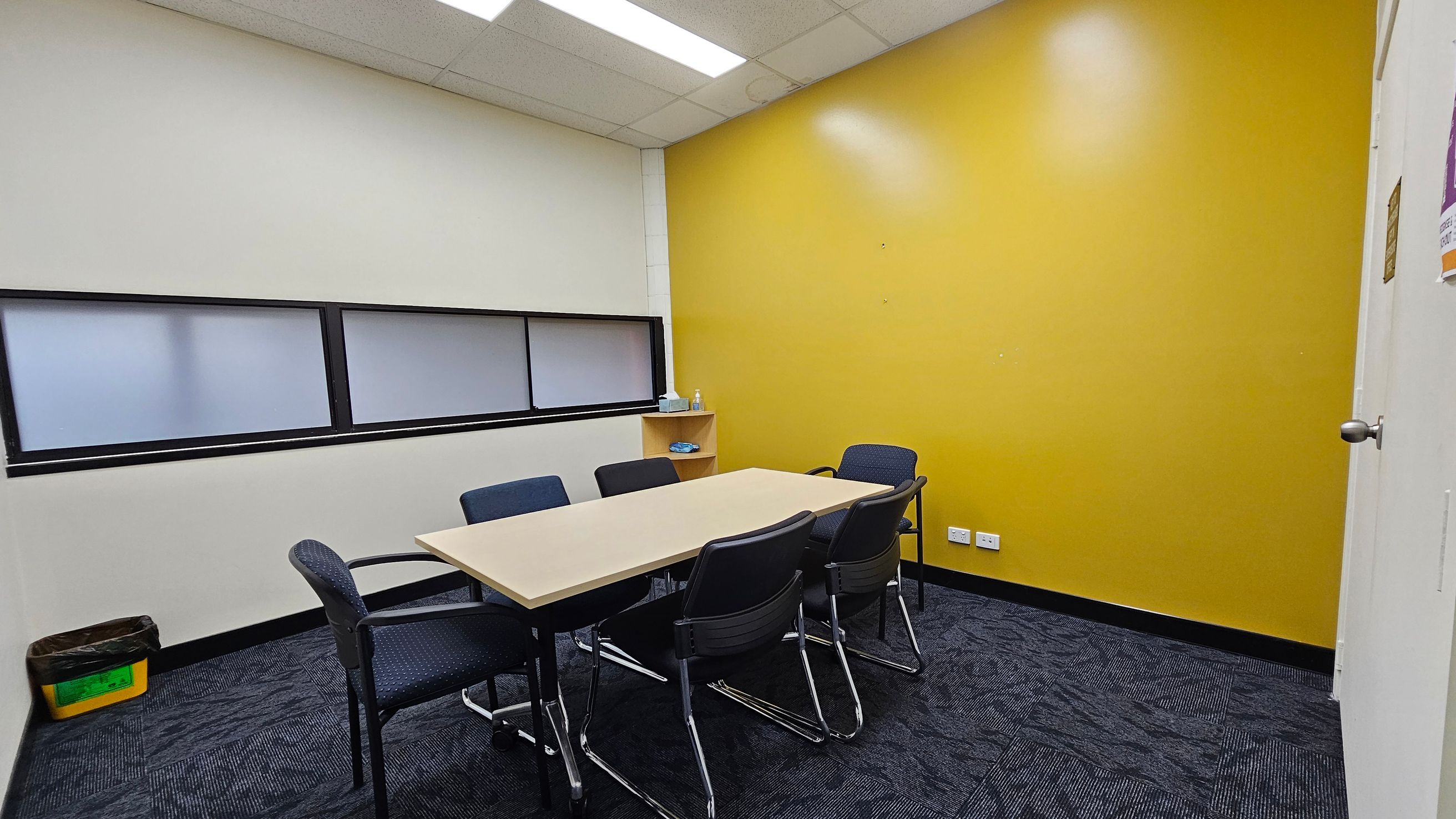 A small meeting room with a rectangular table surrounded by six chairs. The left wall is white with a horizontal frosted window, and a yellow bin is on the floor. The right wall is bright yellow with a small shelf holding a bottle and some items. The floor is covered with dark patterned carpet tiles. The ceiling has white panels with fluorescent lights. A small meeting room with a rectangular table surrounded by six chairs. The left wall is white with a horizontal frosted window, and a yellow bin is on the floor. The right wall is bright yellow with a small shelf holding a bottle and some items. The floor is covered with dark patterned carpet tiles. The ceiling has white panels with fluorescent lights.