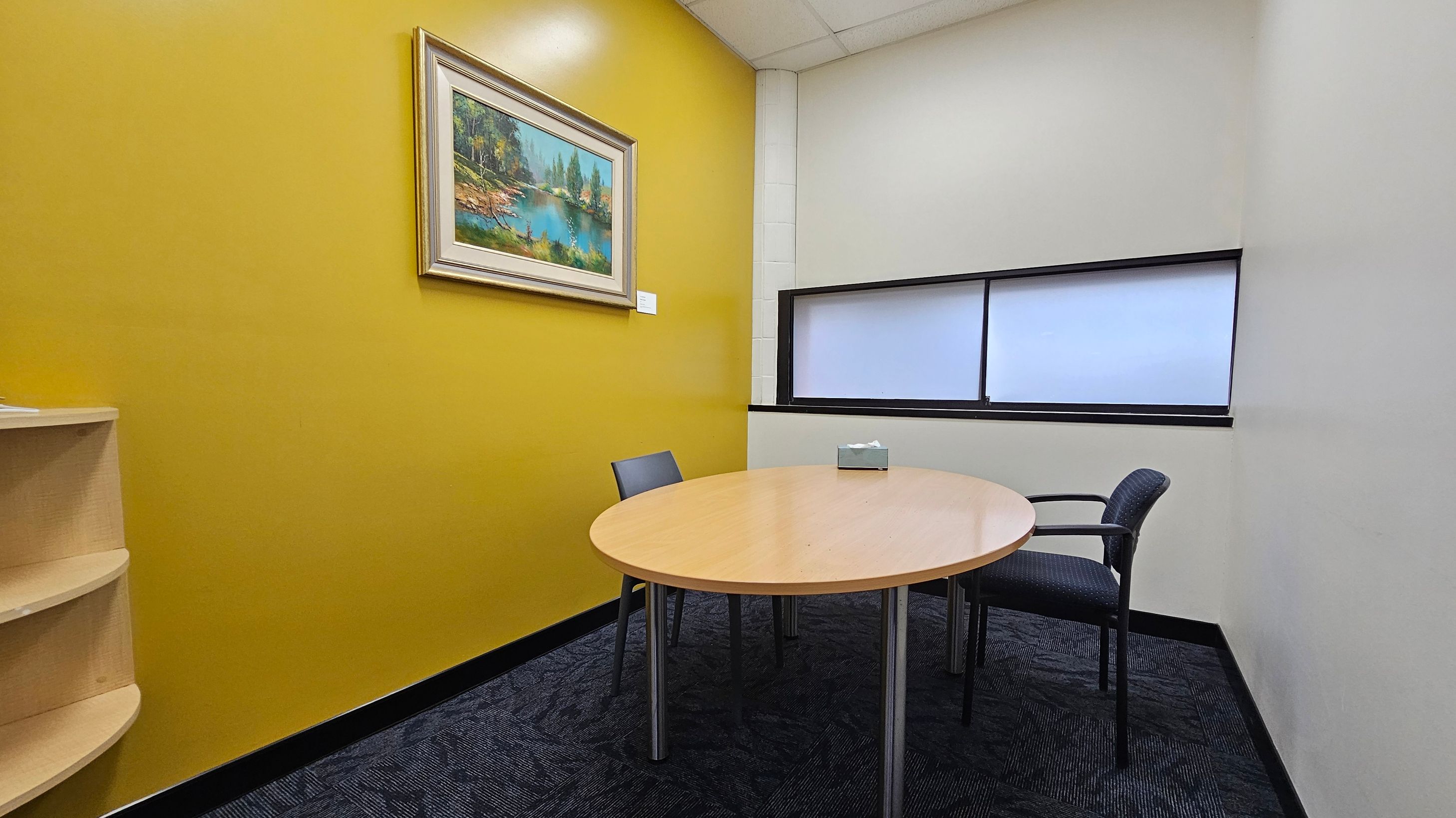 A small office room with a mustard yellow wall on the left, featuring a framed landscape painting. Below the painting is a light wood shelf. In the centre, a round wooden table with a tissue box on top is surrounded by two chairs. The right wall is white with a rectangular window featuring frosted glass. The floor is covered with dark patterned carpet, and the ceiling is white with recessed lighting. A small office room with a mustard yellow wall on the left, featuring a framed landscape painting. Below the painting is a light wood shelf. In the centre, a round wooden table with a tissue box on top is surrounded by two chairs. The right wall is white with a rectangular window featuring frosted glass. The floor is covered with dark patterned carpet, and the ceiling is white with recessed lighting.