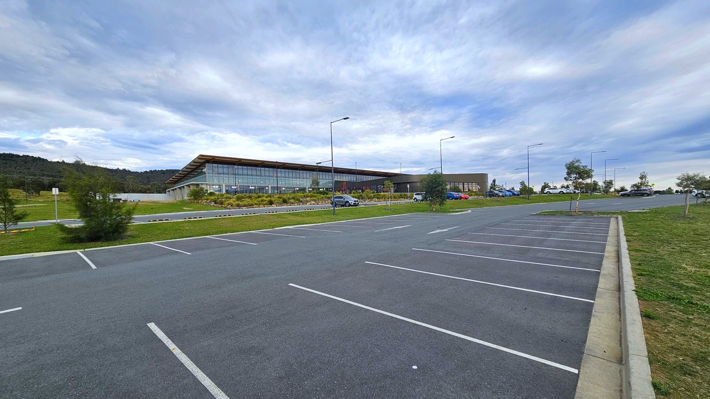 The Stromlo Leisure Centre has a contemporary architectural design incorporating glass, steel, and timber elements. Adjacent to the building, there is ample car parking with a few cars parked. Tall street lights, along with trees and grass, surround the area. The Stromlo Leisure Centre has a contemporary architectural design incorporating glass, steel, and timber elements. Adjacent to the building, there is ample car parking with a few cars parked. Tall street lights, along with trees and grass, surround the area.