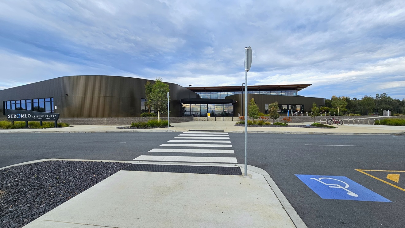 The Stromlo Leisure Centre has a modern architectural design, blending glass, steel, and timber elements. A facility name sign stands in front of the building on the left. On the right, bike racks are available, and adjacent accessible parking is marked with blue wheelchair signs. A pedestrian lane in the middle is marked with truncated domes. A gravel area is situated on the left. Tall street lights, trees, and grass surround the facility. The Stromlo Leisure Centre has a modern architectural design, blending glass, steel, and timber elements. A facility name sign stands in front of the building on the left. On the right, bike racks are available, and adjacent accessible parking is marked with blue wheelchair signs. A pedestrian lane in the middle is marked with truncated domes. A gravel area is situated on the left. Tall street lights, trees, and grass surround the facility.