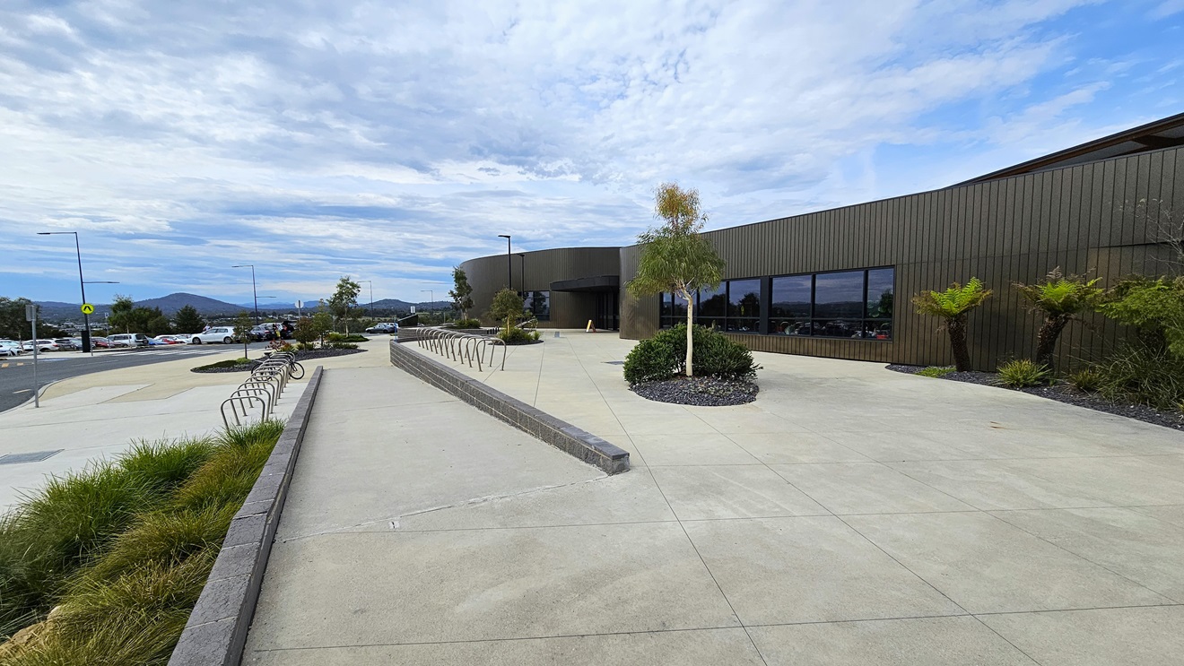The Stromlo Leisure Centre has a contemporary architectural design, blending glass, steel, and timber elements. A ramp entry is located on the left side of the entrance, accompanied by visible bike racks. The expansive concourse is enhanced with a landscape of plants, palm trees, and grass. The Stromlo Leisure Centre has a contemporary architectural design, blending glass, steel, and timber elements. A ramp entry is located on the left side of the entrance, accompanied by visible bike racks. The expansive concourse is enhanced with a landscape of plants, palm trees, and grass.