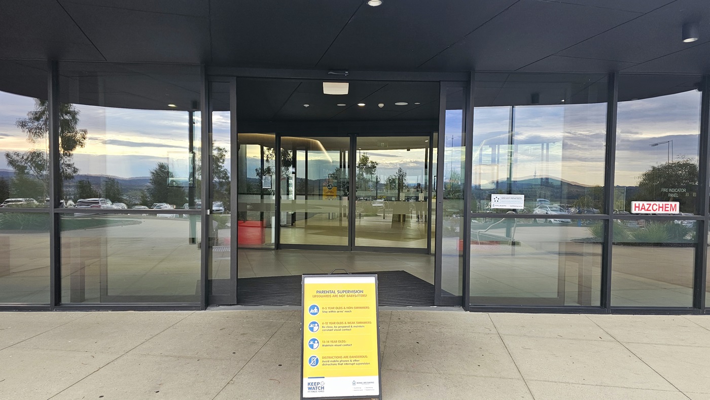 The building entrance features two sets of automatic glass doors with non-slippery mats. A free-standing yellow banner is positioned in front of the entrance. Signs and safety stickers are affixed to the glass wall. The building entrance features two sets of automatic glass doors with non-slippery mats. A free-standing yellow banner is positioned in front of the entrance. Signs and safety stickers are affixed to the glass wall.