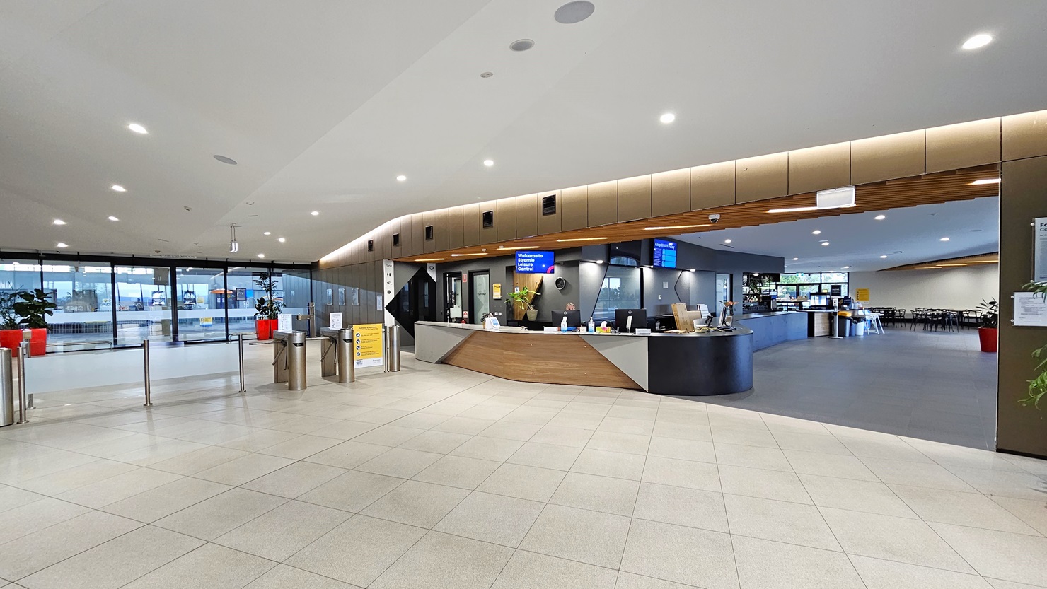 An automatic glass door, adorned with posters, provides access to the facility. Red planter boxes with indoor plants are positioned on both sides. Cushioned benches are on the left. The left wall features a brown wall with a flat-screen TV and retractable banners. To the right is the reception desk, featuring a brown and white front. A flat-screen TV is mounted from the ceiling. In the middle is a barrier gate. The ceiling features a white with scattered LED lights. An automatic glass door, adorned with posters, provides access to the facility. Red planter boxes with indoor plants are positioned on both sides. Cushioned benches are on the left. The left wall features a brown wall with a flat-screen TV and retractable banners. To the right is the reception desk, featuring a brown and white front. A flat-screen TV is mounted from the ceiling. In the middle is a barrier gate. The ceiling features a white with scattered LED lights.