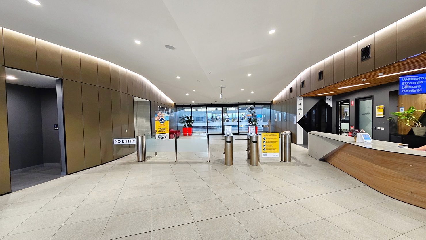 To the right is the reception desk, sporting a brown, white, and black front. A ceiling-mounted flat-screen TV is visible. In the middle is a turnstile gate. Further back, the main entrance is adorned with two red planter boxes and free-standing banners. On the left is an accessible bathroom. The reception desk extends into the corridor, leading to a seating area with chairs and tables on the right. The ceiling features a combination of white and brown with scattered LED lights. To the right is the reception desk, sporting a brown, white, and black front. A ceiling-mounted flat-screen TV is visible. In the middle is a turnstile gate. Further back, the main entrance is adorned with two red planter boxes and free-standing banners. On the left is an accessible bathroom. The reception desk extends into the corridor, leading to a seating area with chairs and tables on the right. The ceiling features a combination of white and brown with scattered LED lights.