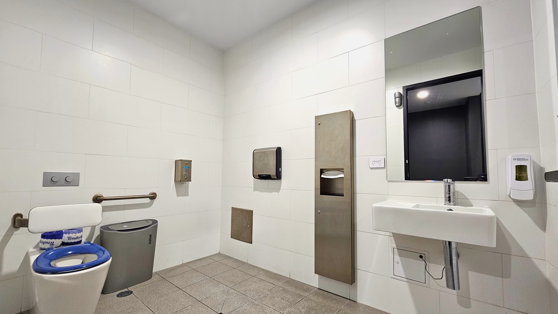 In the accessible bathroom, a toilet bowl with handrails is situated to the right, accompanied by a nearby rubbish bin. Adjacent to it, on the right, there is a wash basin, a mirror, and a soap dispenser. Additional features include a tissue dispenser and a hand dryer. The floor is covered in grey tiles, while the ceiling and walls are uniformly white. In the accessible bathroom, a toilet bowl with handrails is situated to the right, accompanied by a nearby rubbish bin. Adjacent to it, on the right, there is a wash basin, a mirror, and a soap dispenser. Additional features include a tissue dispenser and a hand dryer. The floor is covered in grey tiles, while the ceiling and walls are uniformly white.