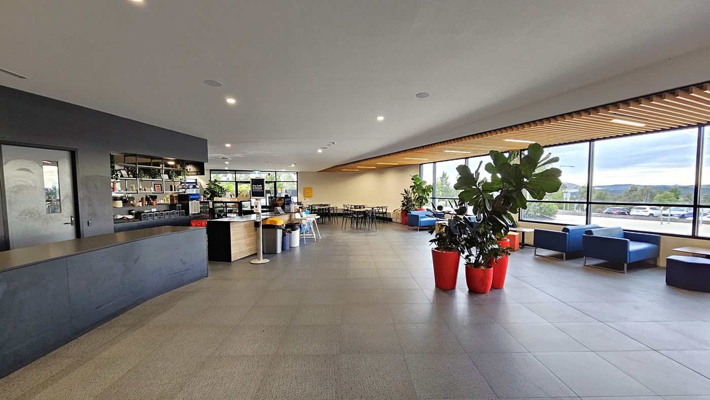 To the left is the extended reception desk and the café, featuring a sanitizer in the centre. The face exhibits a brown timber design. In the middle, there are pots of indoor plants. On the right, couches are positioned near the glass wall, and further in, there are movable plastic chairs and tables. Wooden planks adorn the ceiling on the right. The floor is white with LED lights, and grey tiles cover the floor. To the left is the extended reception desk and the café, featuring a sanitizer in the centre. The face exhibits a brown timber design. In the middle, there are pots of indoor plants. On the right, couches are positioned near the glass wall, and further in, there are movable plastic chairs and tables. Wooden planks adorn the ceiling on the right. The floor is white with LED lights, and grey tiles cover the floor.