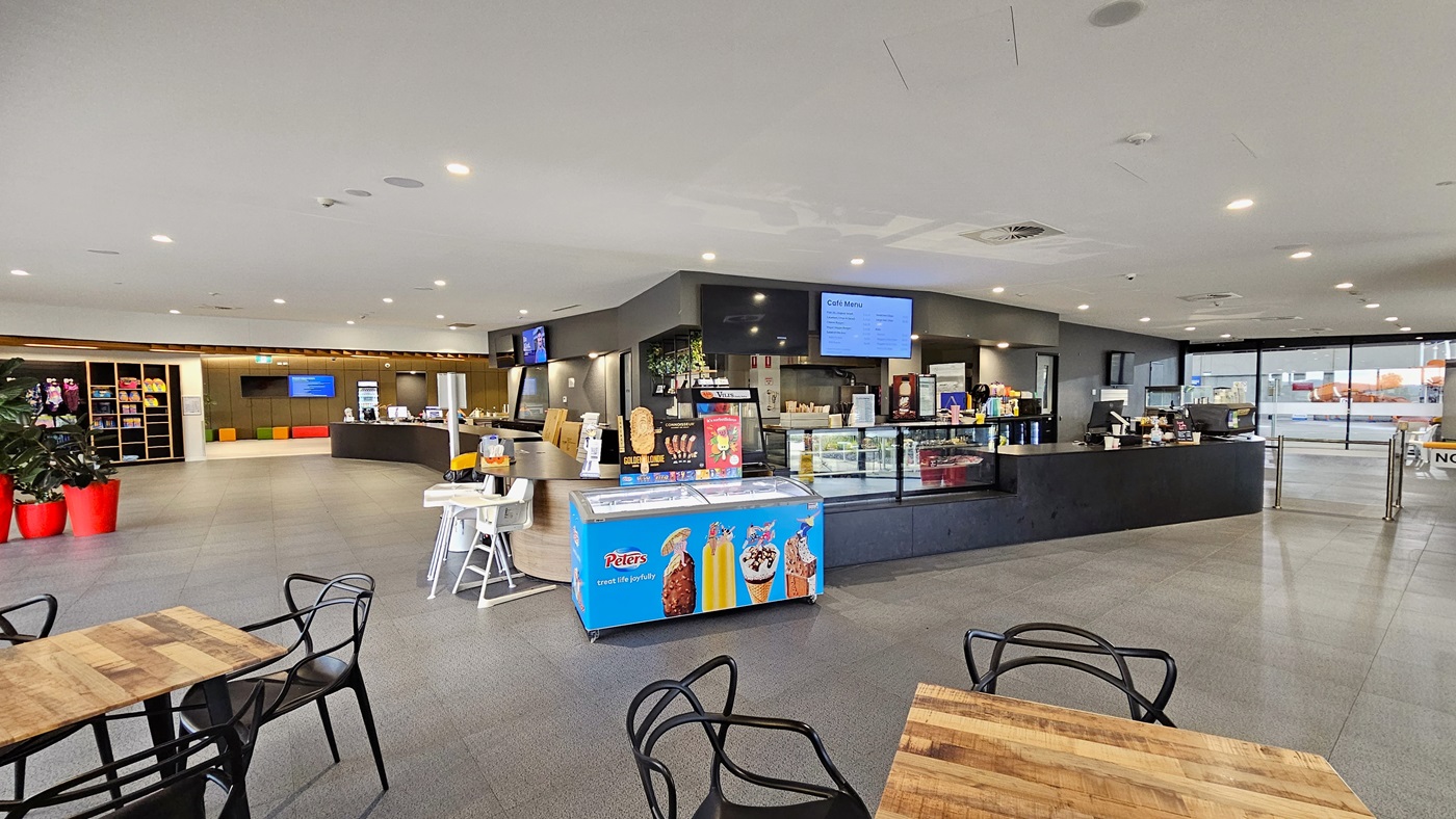 In the centre is the café with a black counter desk and a glass food display rack. A flat-screen TV is mounted on the wall, and an ice cream freezer is placed in the corner. On the left, a table holding flyers is situated, and further left, indoor plants in bright red pots are displayed along with a storage rack showcasing retail items. Movable plastic chairs and tables are arranged in front of the café. To the right is a turnstile gate. In the centre is the café with a black counter desk and a glass food display rack. A flat-screen TV is mounted on the wall, and an ice cream freezer is placed in the corner. On the left, a table holding flyers is situated, and further left, indoor plants in bright red pots are displayed along with a storage rack showcasing retail items. Movable plastic chairs and tables are arranged in front of the café. To the right is a turnstile gate.