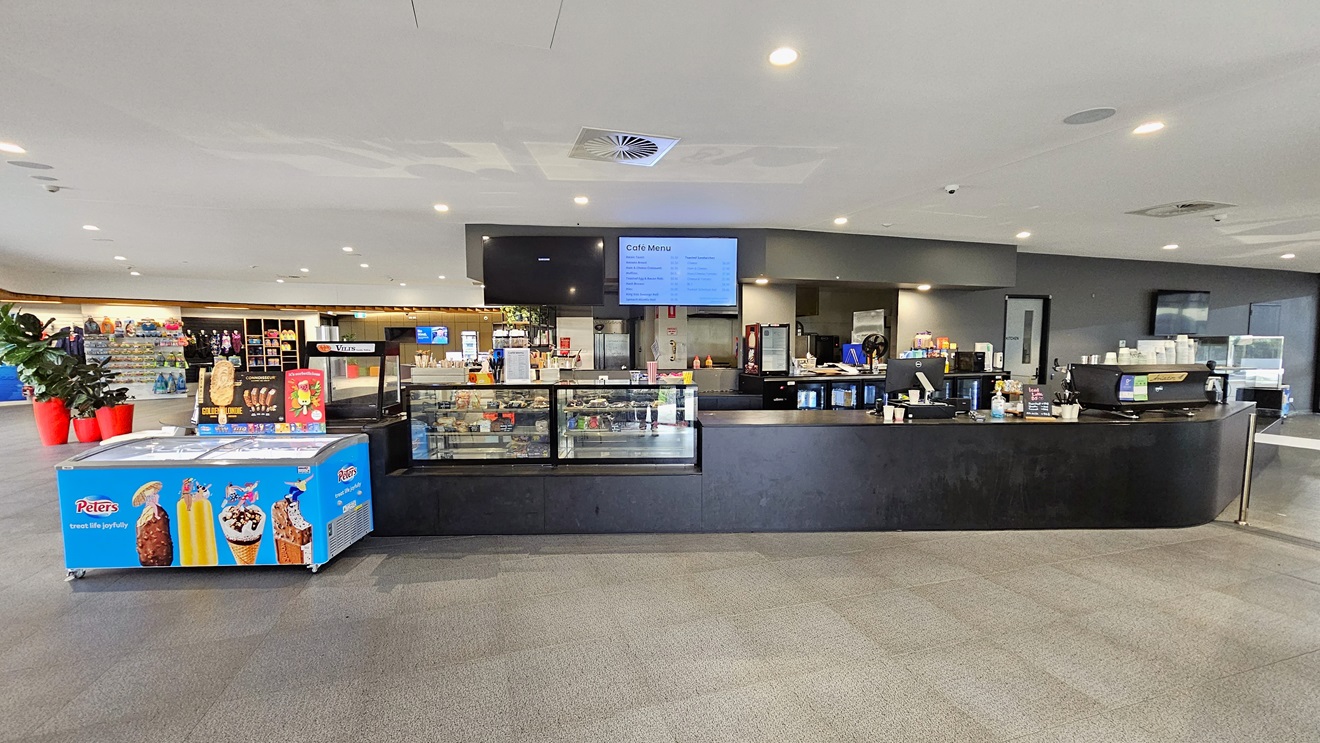 In the centre is the café with a black counter desk and a glass food display rack. A flat-screen TV is mounted on the wall, and an ice cream freezer is placed in the corner. On the left, a table holding flyers is situated, and further left, indoor plants in bright red pots are displayed along with a storage rack showcasing retail items. To the right is a turnstile gate. The floor is white with LED lights, and grey tiles cover the floor. In the centre is the café with a black counter desk and a glass food display rack. A flat-screen TV is mounted on the wall, and an ice cream freezer is placed in the corner. On the left, a table holding flyers is situated, and further left, indoor plants in bright red pots are displayed along with a storage rack showcasing retail items. To the right is a turnstile gate. The floor is white with LED lights, and grey tiles cover the floor.