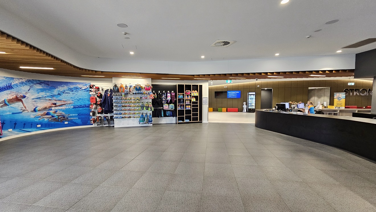 In the middle is a retail area displaying goggles, swimwear, and swimming accessories. To the right is the reception desk featuring an extensive black desk. To the left, the wall is adorned with a life-size poster of swimmers underwater. The ceiling has a cove design with wooden plank accents and LED lights. Grey tiles cover the floor. Further left, the brown wall holds flat-screen TVs, banners, and the facility name. In the middle is a retail area displaying goggles, swimwear, and swimming accessories. To the right is the reception desk featuring an extensive black desk. To the left, the wall is adorned with a life-size poster of swimmers underwater. The ceiling has a cove design with wooden plank accents and LED lights. Grey tiles cover the floor. Further left, the brown wall holds flat-screen TVs, banners, and the facility name.