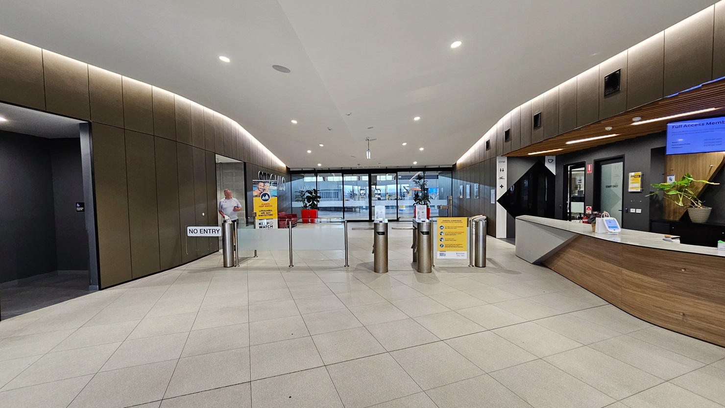 To the right is the reception desk, sporting a brown, white, and black front. A ceiling-mounted flat-screen TV is visible. In the middle is a turnstile gate. Further back, the main entrance is adorned with two red planter boxes and free-standing banners. On the left is an accessible bathroom. The reception desk extends into the corridor, leading to a seating area with chairs and tables on the right. The ceiling features a combination of white and brown with scattered LED lights. To the right is the reception desk, sporting a brown, white, and black front. A ceiling-mounted flat-screen TV is visible. In the middle is a turnstile gate. Further back, the main entrance is adorned with two red planter boxes and free-standing banners. On the left is an accessible bathroom. The reception desk extends into the corridor, leading to a seating area with chairs and tables on the right. The ceiling features a combination of white and brown with scattered LED lights.
