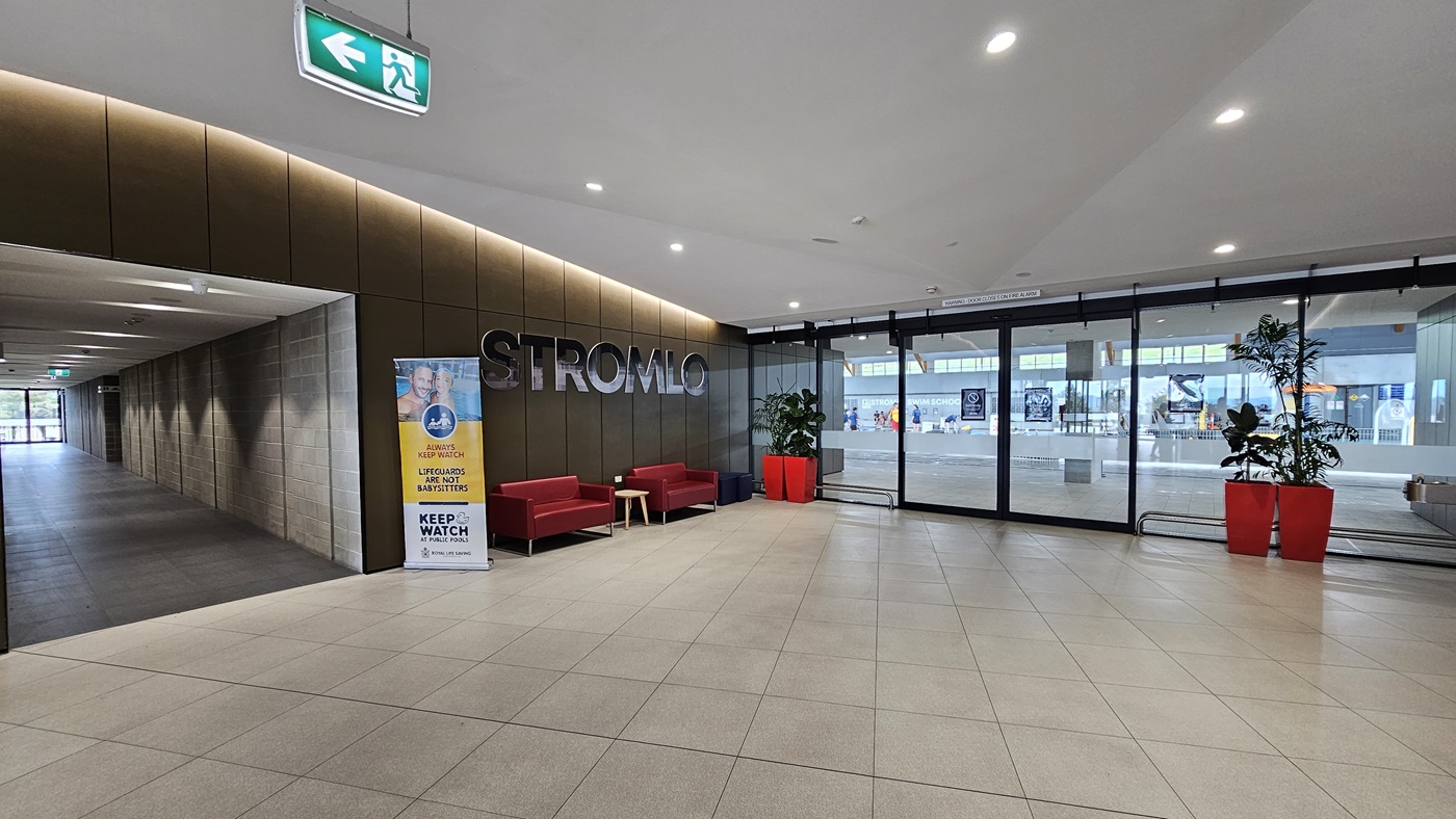 To the left is a corridor with grey brick walls. In the middle, red couches are arranged against the brown wall, with the facility's name on top. A retractable banner is positioned next to it. To the right, a glass door entry equipped with banners serves as the entry to the aquatic facility. Large red planter boxes with indoor plants are placed against the wall. The floor is white with LED lights, and grey tiles cover the floor. To the left is a corridor with grey brick walls. In the middle, red couches are arranged against the brown wall, with the facility's name on top. A retractable banner is positioned next to it. To the right, a glass door entry equipped with banners serves as the entry to the aquatic facility. Large red planter boxes with indoor plants are placed against the wall. The floor is white with LED lights, and grey tiles cover the floor.