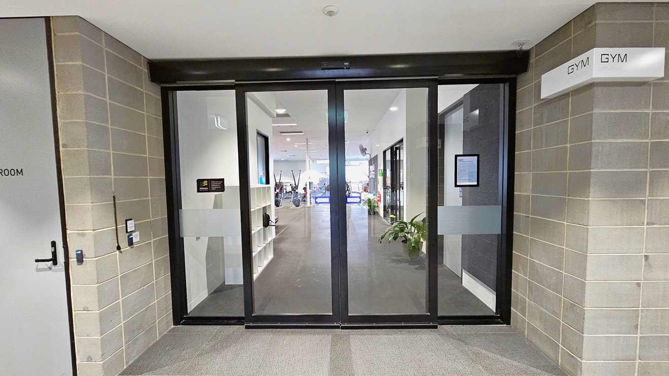 The entrance to the Gymnasium is through an automated glass sliding door adorned with posters and a sticker accent. A “Gym” sign is affixed to the upper left wall. The exterior wall is grey bricks, and the floor is tiled in grey. A grey door is visible on the left. The ceiling is uniformly white. Inside, a storage rack is located near the door. The entrance to the Gymnasium is through an automated glass sliding door adorned with posters and a sticker accent. A “Gym” sign is affixed to the upper left wall. The exterior wall is grey bricks, and the floor is tiled in grey. A grey door is visible on the left. The ceiling is uniformly white. Inside, a storage rack is located near the door.