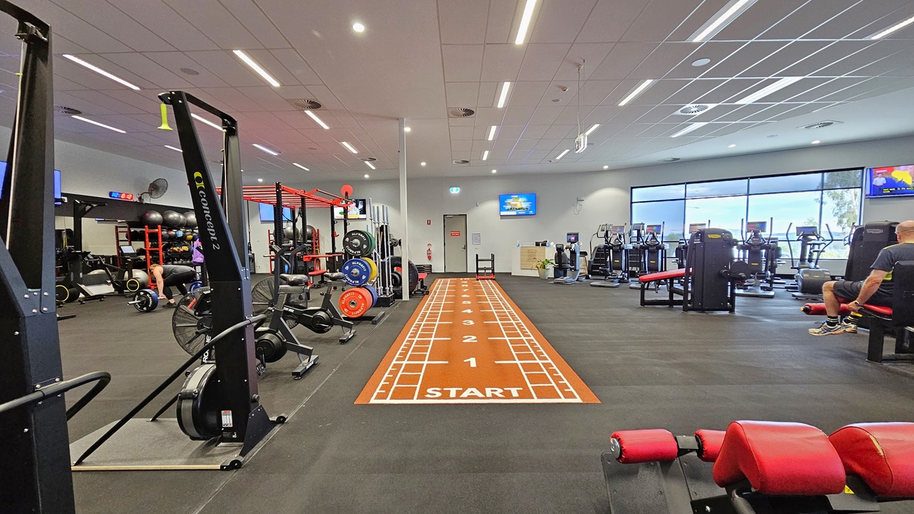 Inside the Gym, the left side houses Smith Machines, SkiErgs, Spin Machines, and free weights. In the middle, there is an orange sprint floor. Additional machines on the right include bench press, stair mill, and treadmills. Flatscreen TVs are mounted on the walls, and a glass wall surrounds the right side. The floor has black rubber tiles, and the ceiling features a white porous design with air conditioning and fluorescent lights. Inside the Gym, the left side houses Smith Machines, SkiErgs, Spin Machines, and free weights. In the middle, there is an orange sprint floor. Additional machines on the right include bench press, stair mill, and treadmills. Flatscreen TVs are mounted on the walls, and a glass wall surrounds the right side. The floor has black rubber tiles, and the ceiling features a white porous design with air conditioning and fluorescent lights.