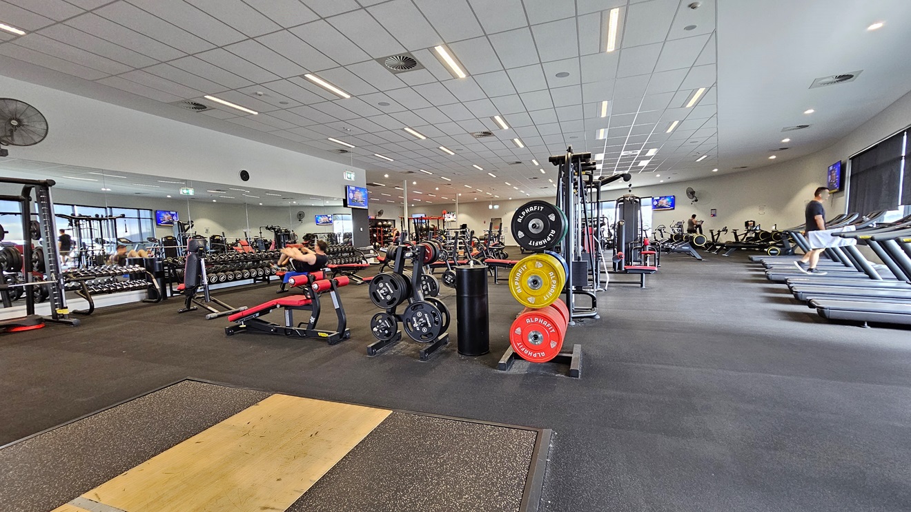 Inside the Gym, the free weights section and a Smith Machine are on the right. In the middle, there's a bench press, incline press, and Lat Pulldown machines. On the right, you'll find treadmills and stationary bikes, with additional equipment positioned behind. The floor is covered with black rubber tiles, with a brown accent and the ceiling features a white porous design with air conditioning and fluorescent lights. Inside the Gym, the free weights section and a Smith Machine are on the right. In the middle, there's a bench press, incline press, and Lat Pulldown machines. On the right, you'll find treadmills and stationary bikes, with additional equipment positioned behind. The floor is covered with black rubber tiles, with a brown accent and the ceiling features a white porous design with air conditioning and fluorescent lights.